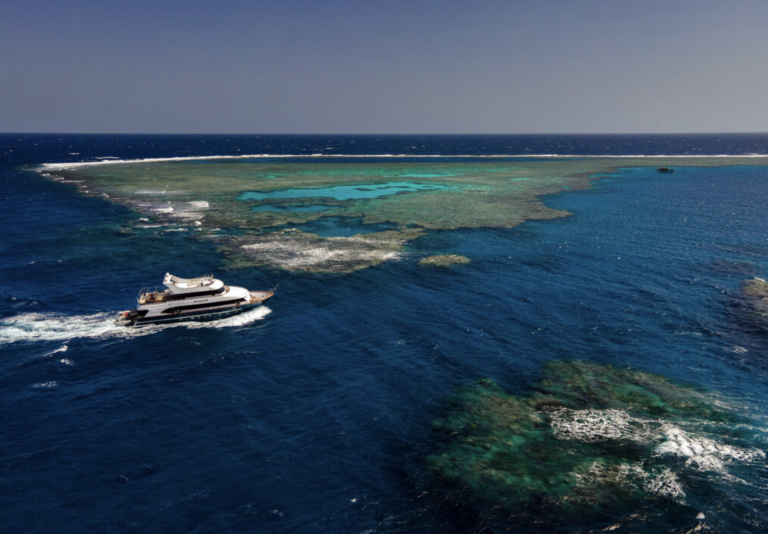 A yacht sailing near a coral reef in the ocean under a clear sky.