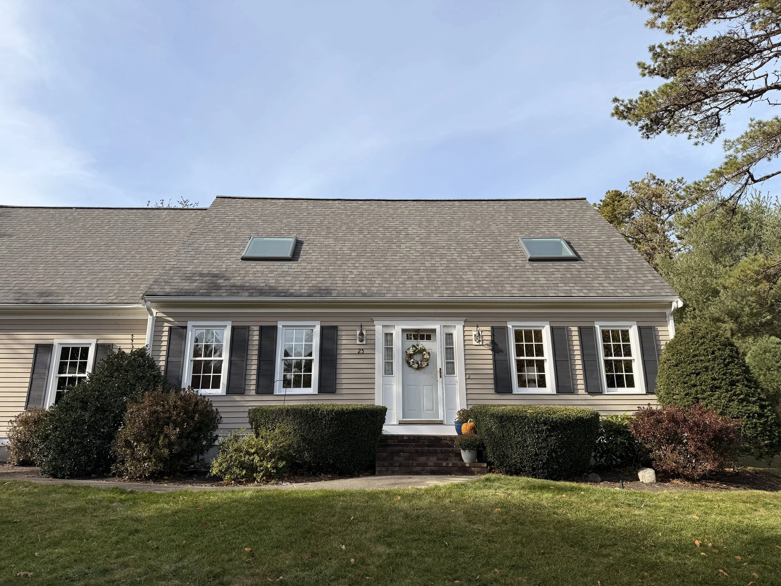 Front view of a house with beige siding, black shutters, two skylights on the roof, and a white front door decorated with a wreath, flanked by two wall-mounted lanterns, and surrounded by landscaped bushes and trees.