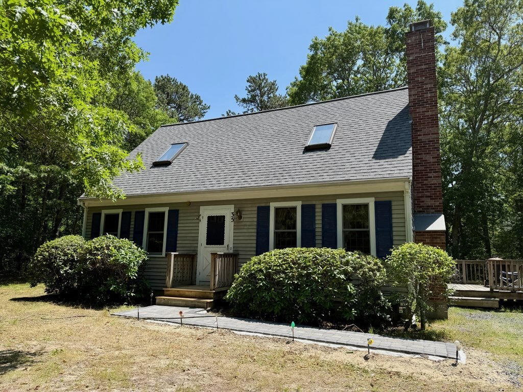 A two-story house with a steep gray roof, blue shutters, and a chimney, surrounded by green trees and shrubs, with a small walkway leading to the front door.