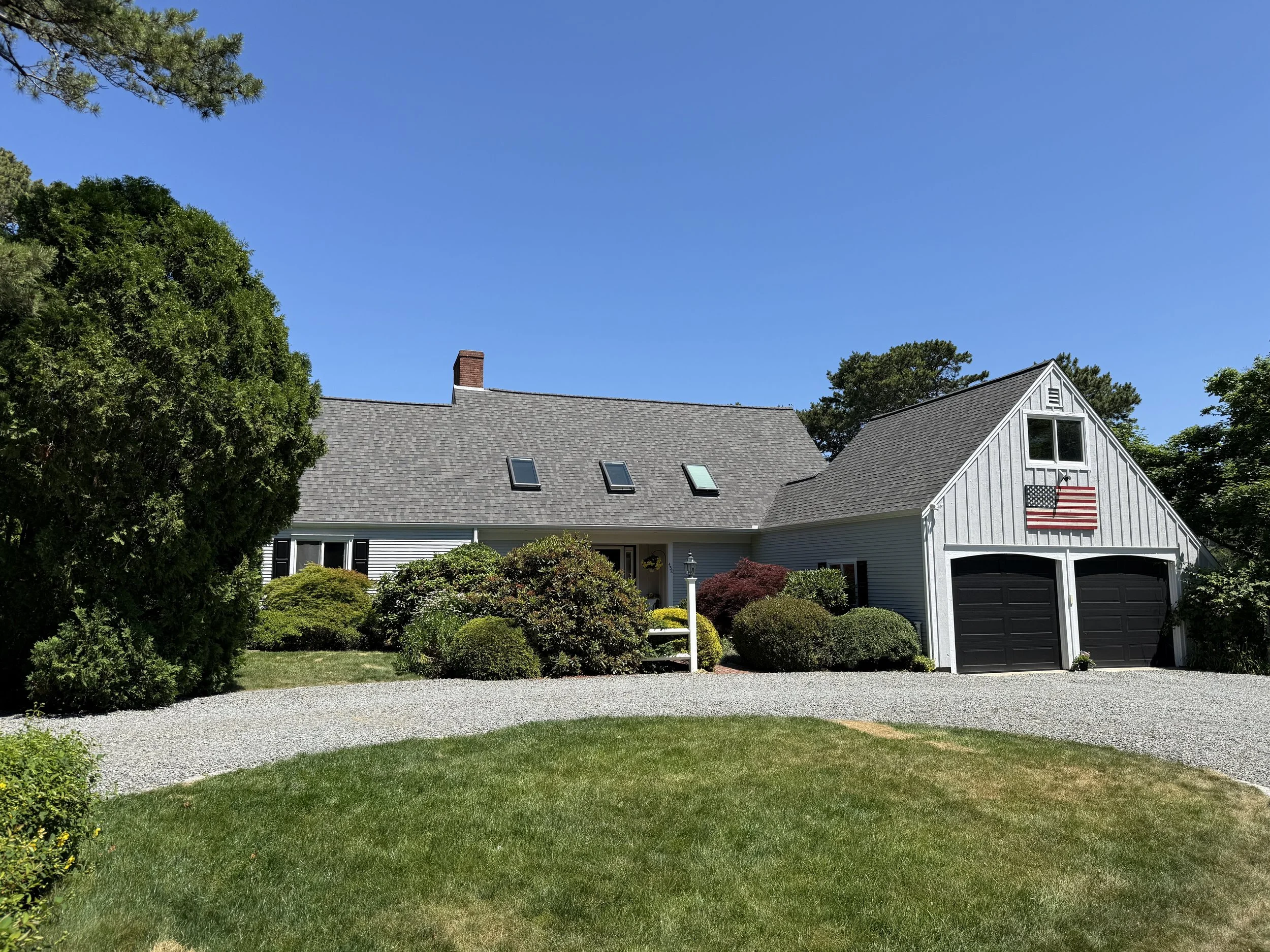 A house with white siding, a gray shingled roof, and a garage with two black doors. An American flag hangs above the garage. The front yard has green grass, bushes, and trees under a blue sky.
