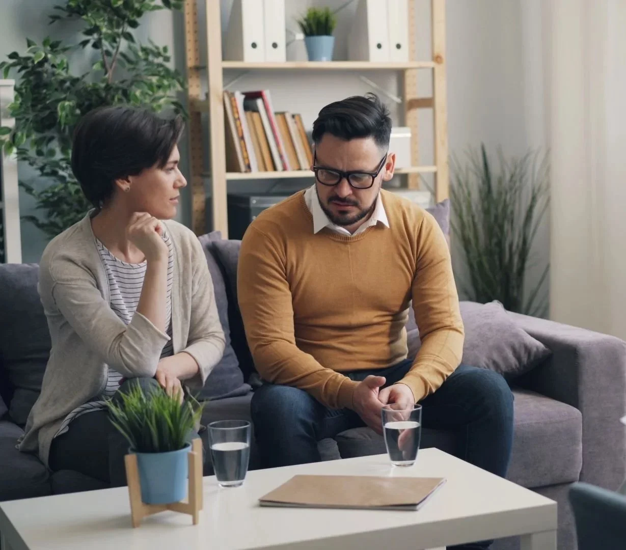 A man and woman sitting on a gray couch in a living room, having a serious conversation. The woman has short dark hair and is wearing a beige cardigan, while the man has dark hair, glasses, and is wearing a mustard sweater. They have glasses of water on the white coffee table in front of them, which also has a brown folder and a small potted plant.