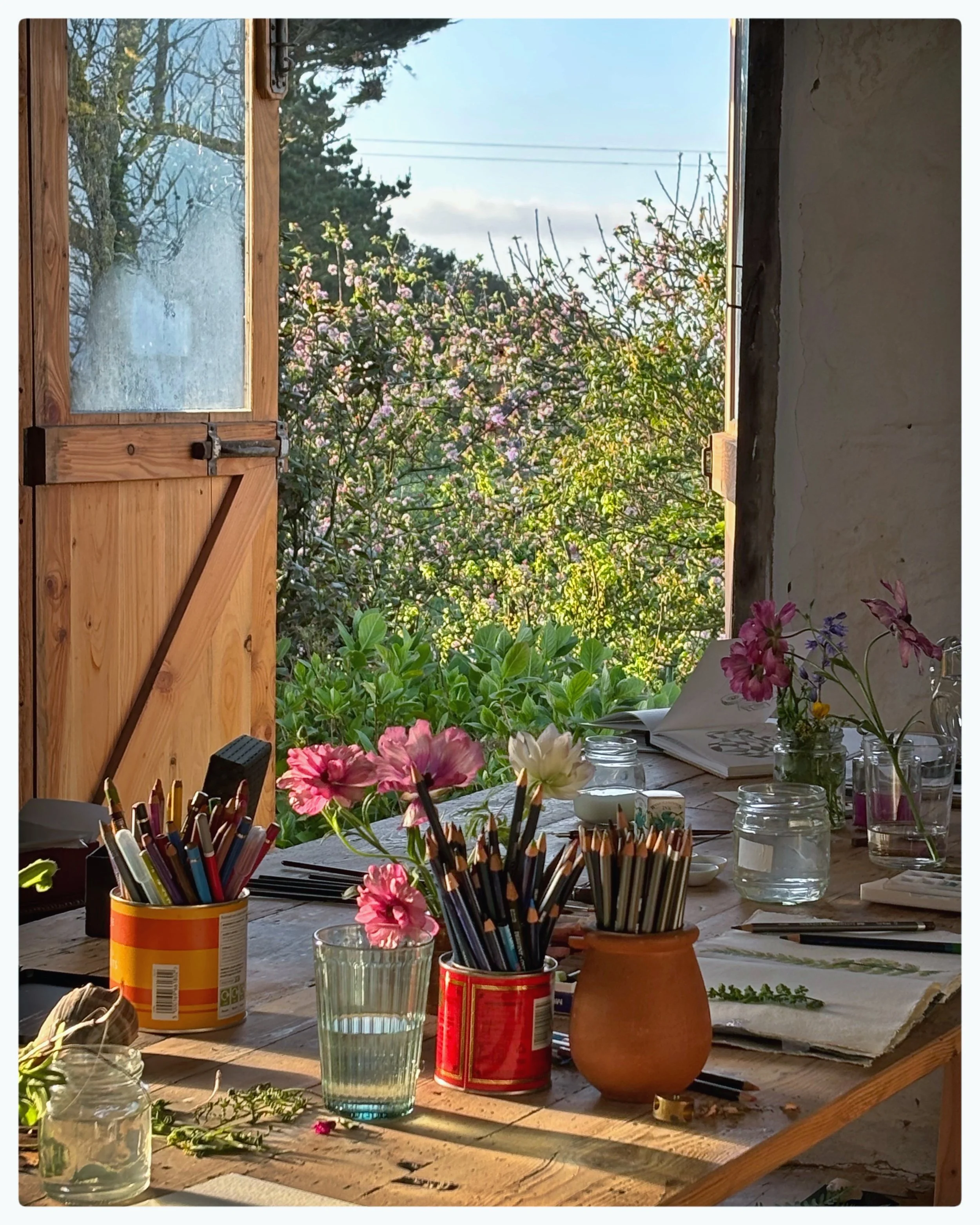 A rustic wooden table outdoors with art supplies, glass jars, and fresh flowers, overlooking blooming trees and a blue sky through an open window.