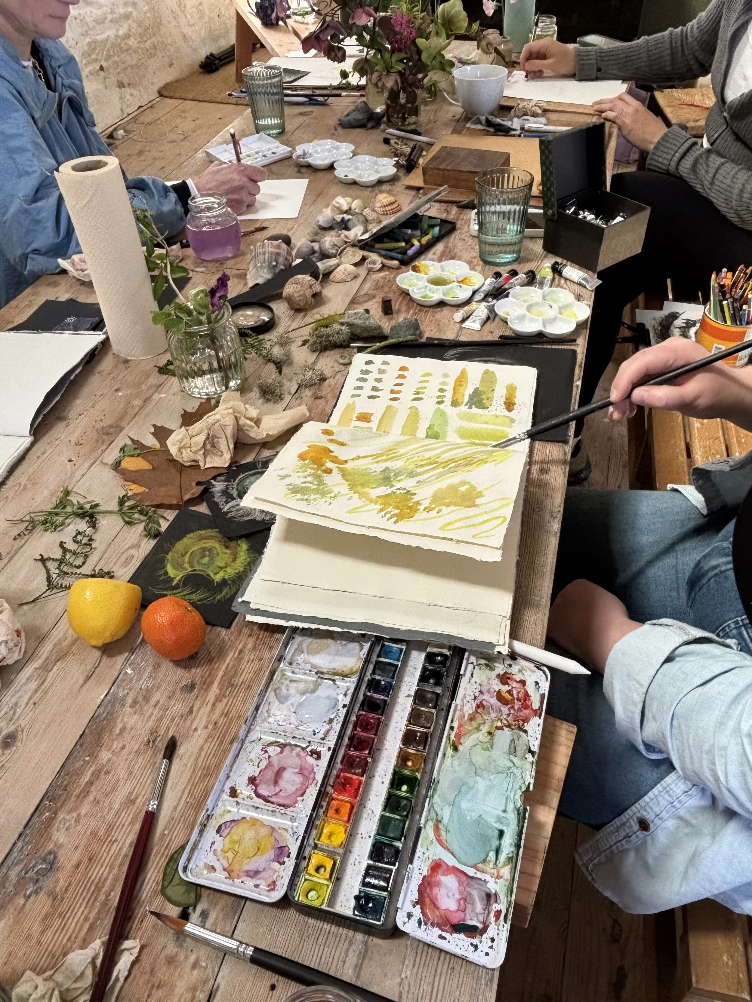A group of people at a table engaged in painting and art activities, with various art supplies, paints, brushes, fruits, and natural objects scattered on the table.