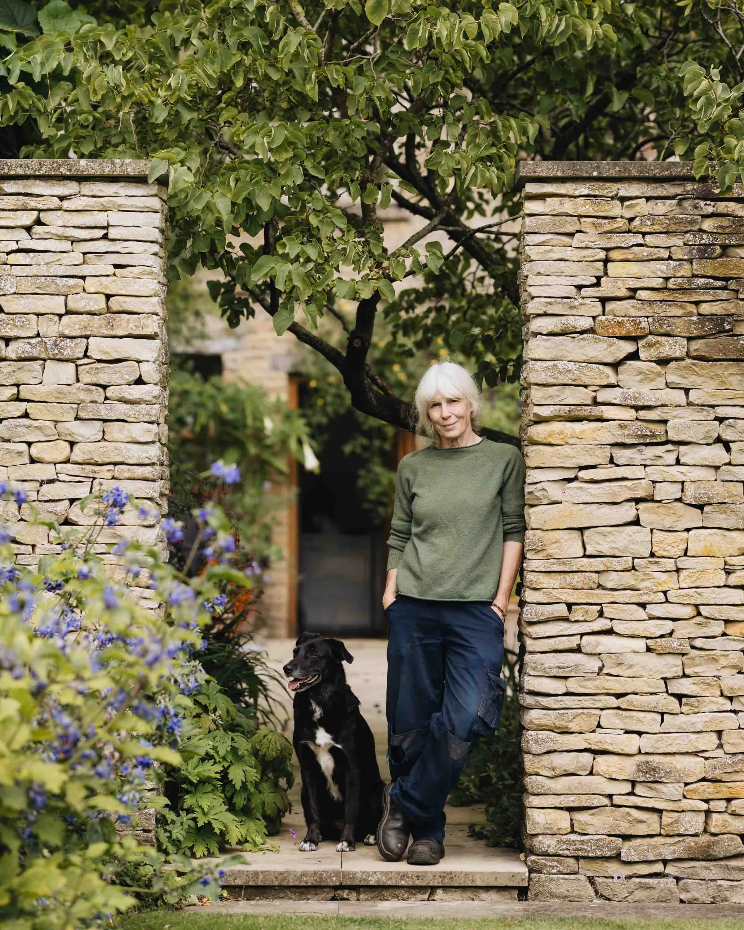 A woman with white hair stands on a stone porch with her hand in her pocket, next to a black dog, in front of a stone wall and a tree with green leaves, with purple flowers nearby.