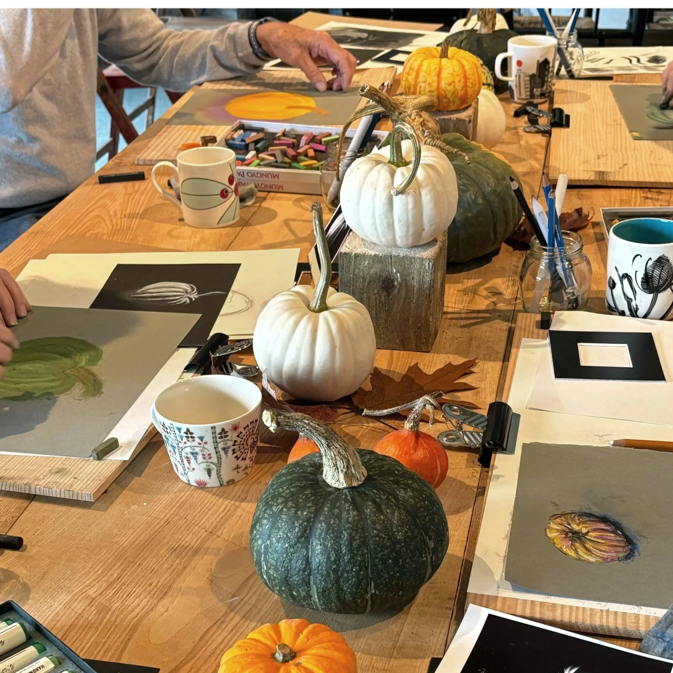 Table decorated with white, green, and orange pumpkins, fall leaves, art supplies, and coffee mugs during an art class.