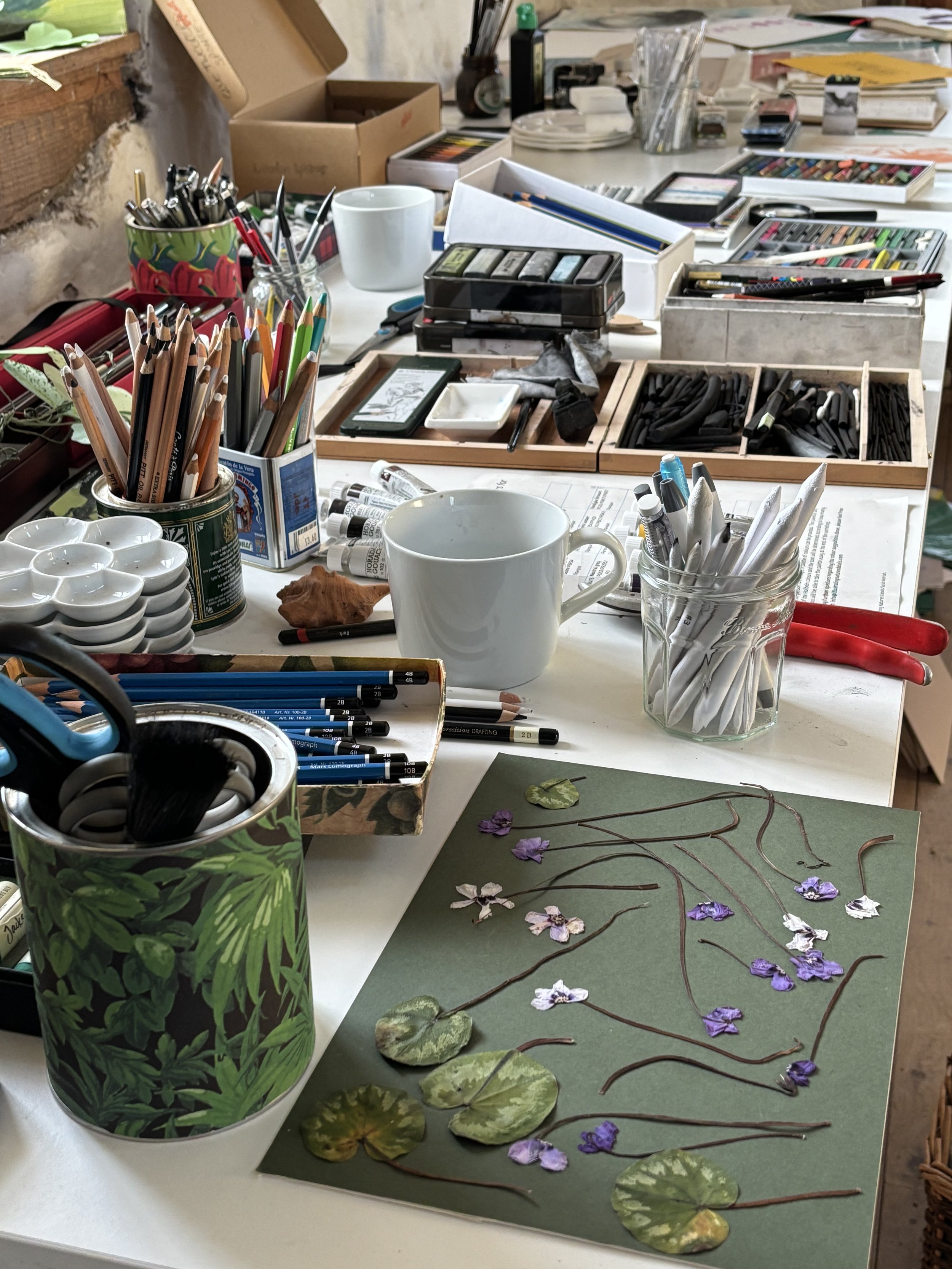 An artist's workspace with various art supplies, including colored pencils, markers, paint tubes, brushes, and paper. There are also cups, boxes, and a floral-themed container, along with a collage project featuring pressed leaves and flowers on gree