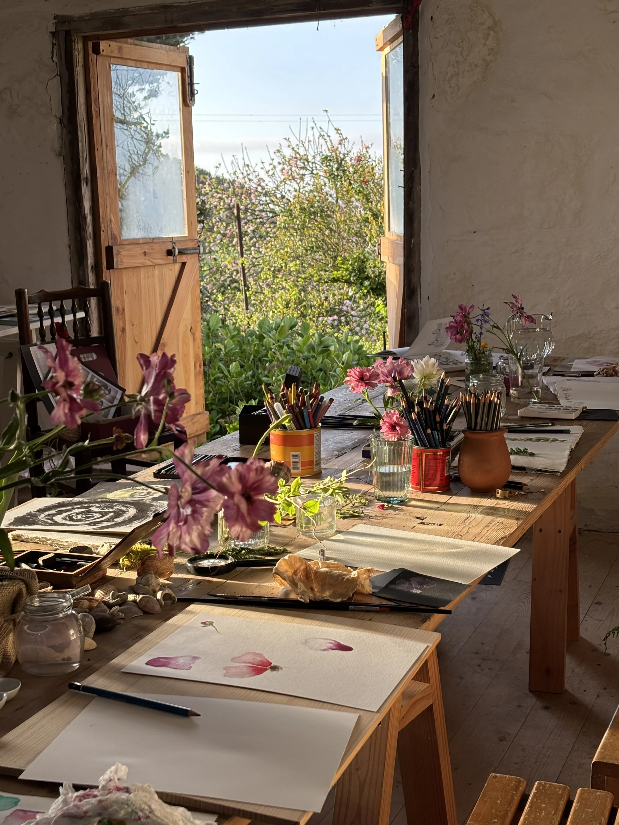 A rustic art studio with a large wooden table filled with watercolor paintings, brushes, jars, and flowers, overlooking a garden through open wooden window shutters.