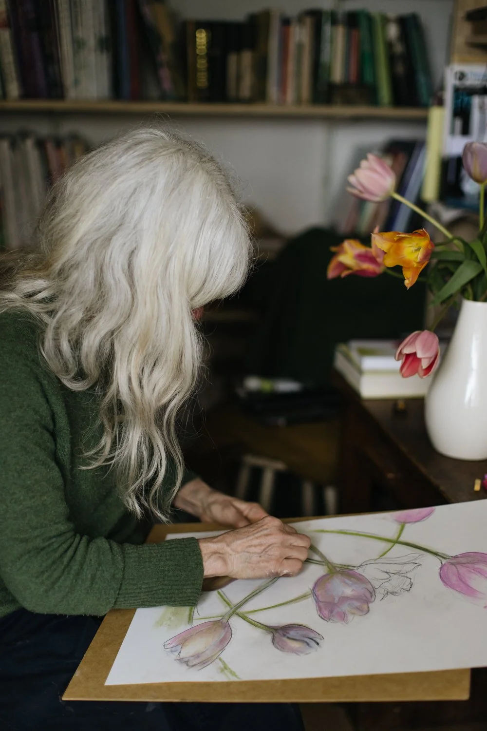 An elderly woman with long, wavy gray hair is working on a botanical watercolor drawing of tulip flowers on a piece of paper placed on a wooden table, with a vase of mixed tulips nearby.