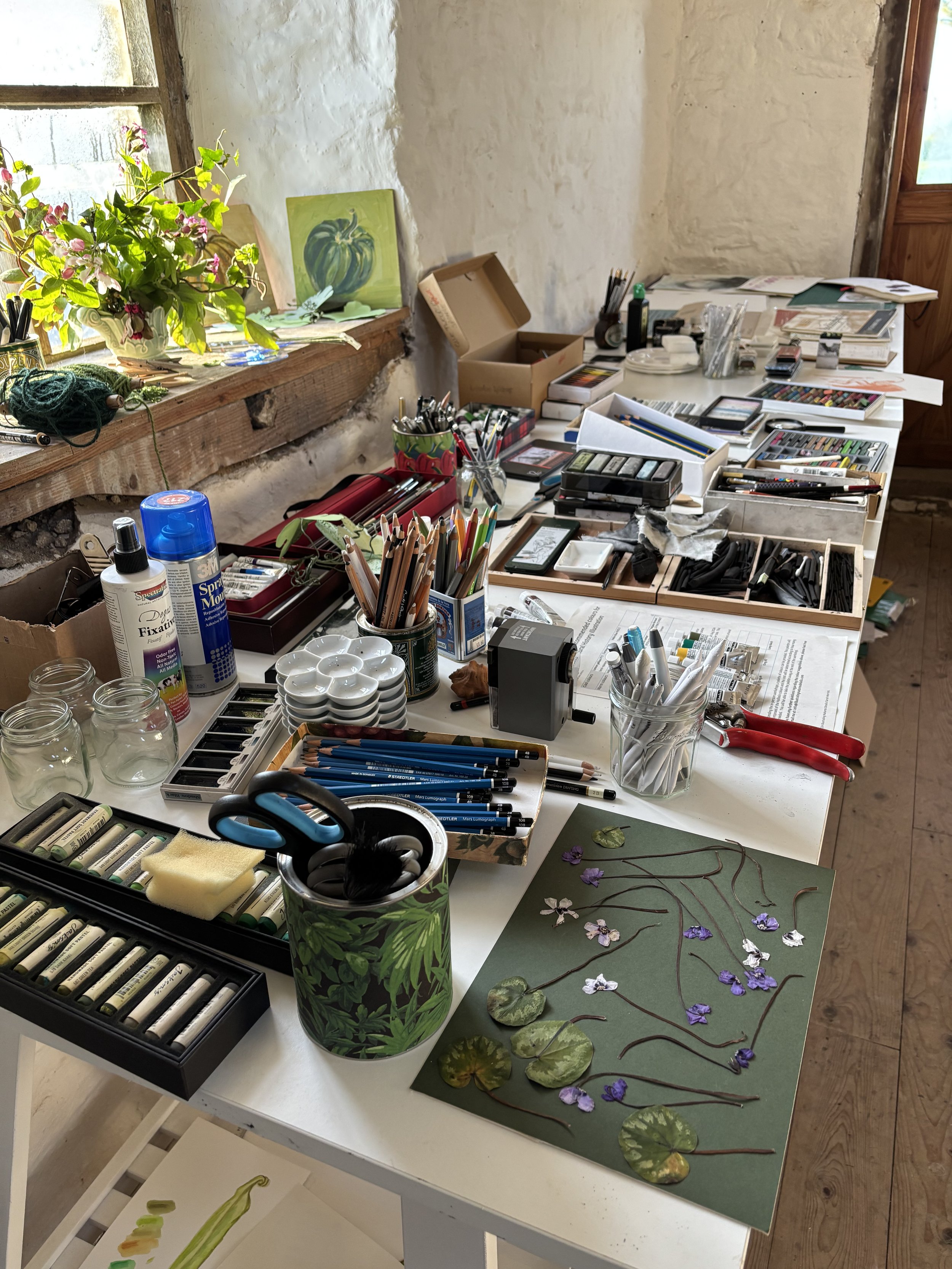 A cluttered art workspace with various art supplies, pencils, paints, scissors, and a floral project on the table. A window with sunlight streams into the room.