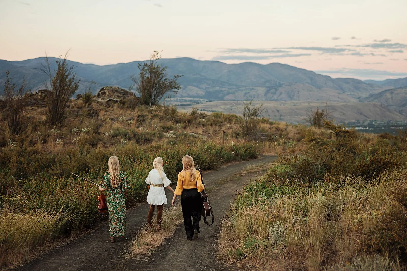 The Gothard Sisters walk down a road in the mountains together, holding instruments. Photo by Ruth H Photography from the album shoot for "Moment in Time" in 2025.