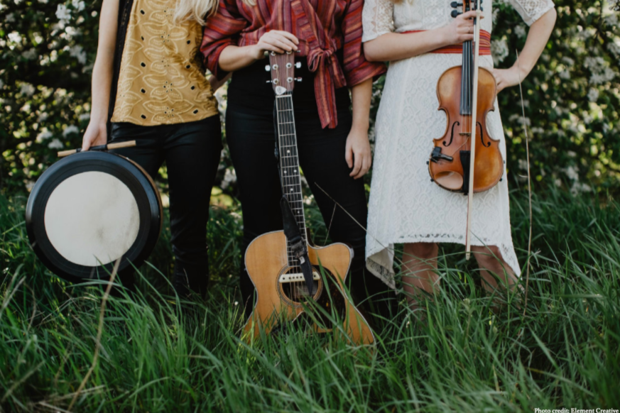 The Gothard Sisters stand in front of a flowering tree with their instruments, photo from 2018. No faces. Solana (left) holds a bodhran. Greta (center) holds a guitar. Willow (right) holds a violin. Photo by Element Creative. Midnight Sun album.