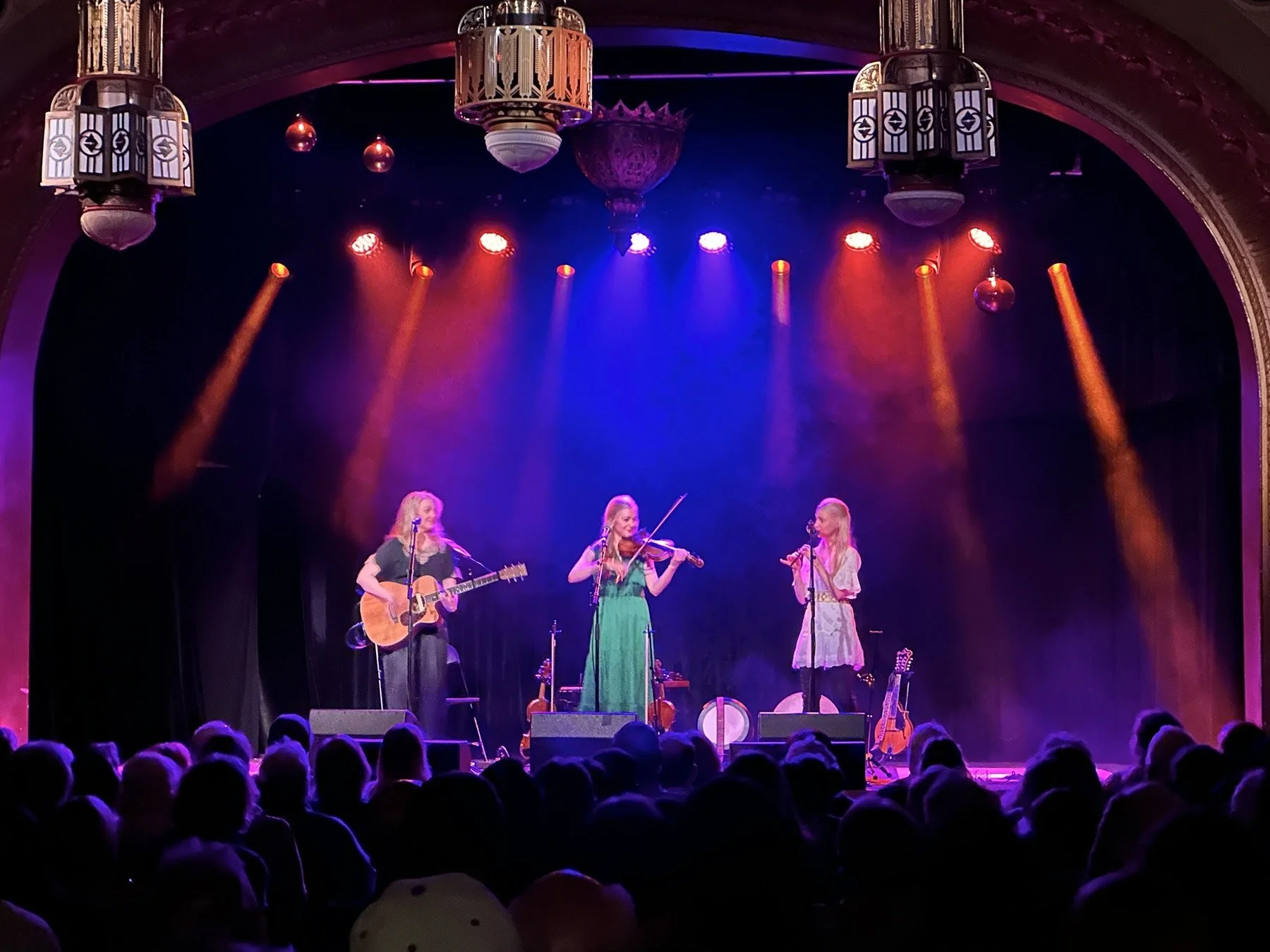 The Gothard Sisters perform onstage at the Spanish Ballroom in Tacoma, WA in March 2026. Live concert. Greta plays guitar, Willow plays violin, Solana plays pennywhistle. An audience watches on.
