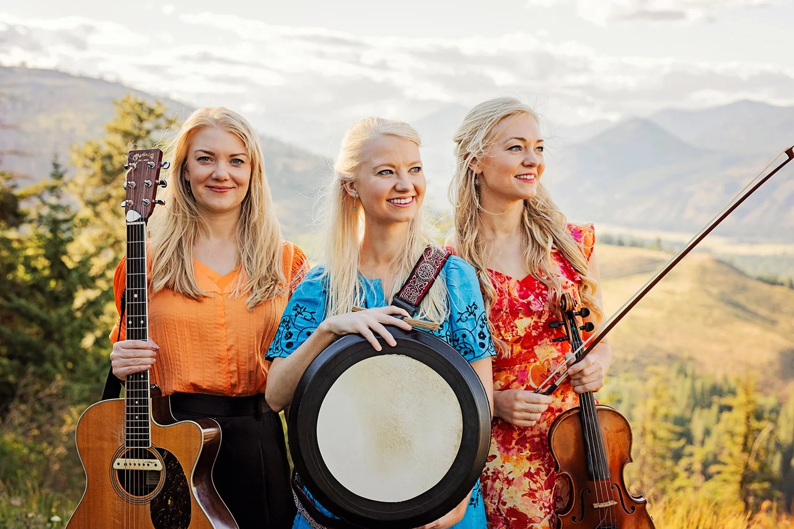The Gothard Sisters, a Celtic Folk trio from the Pacific Northwest, USA, stand on a mountain top holding a guitar, a bodhran and a violin.