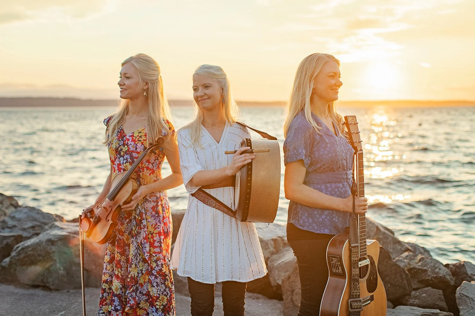 The Gothard Sisters in front of the sunset in their hometown of Edmonds, Washington in Spring 2022. Willow (left) holds a violin. Solana (center) holds a bodhran. Greta (right) holds a guitar. Photo by Ruth H Photography.