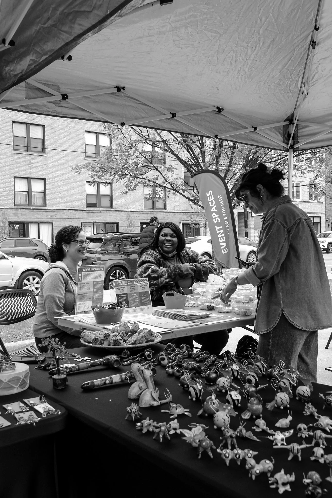 Two people sitting at a table and one person standing, browsing the items on the table at our first ever Mutual Aid Fair event in May 2025.