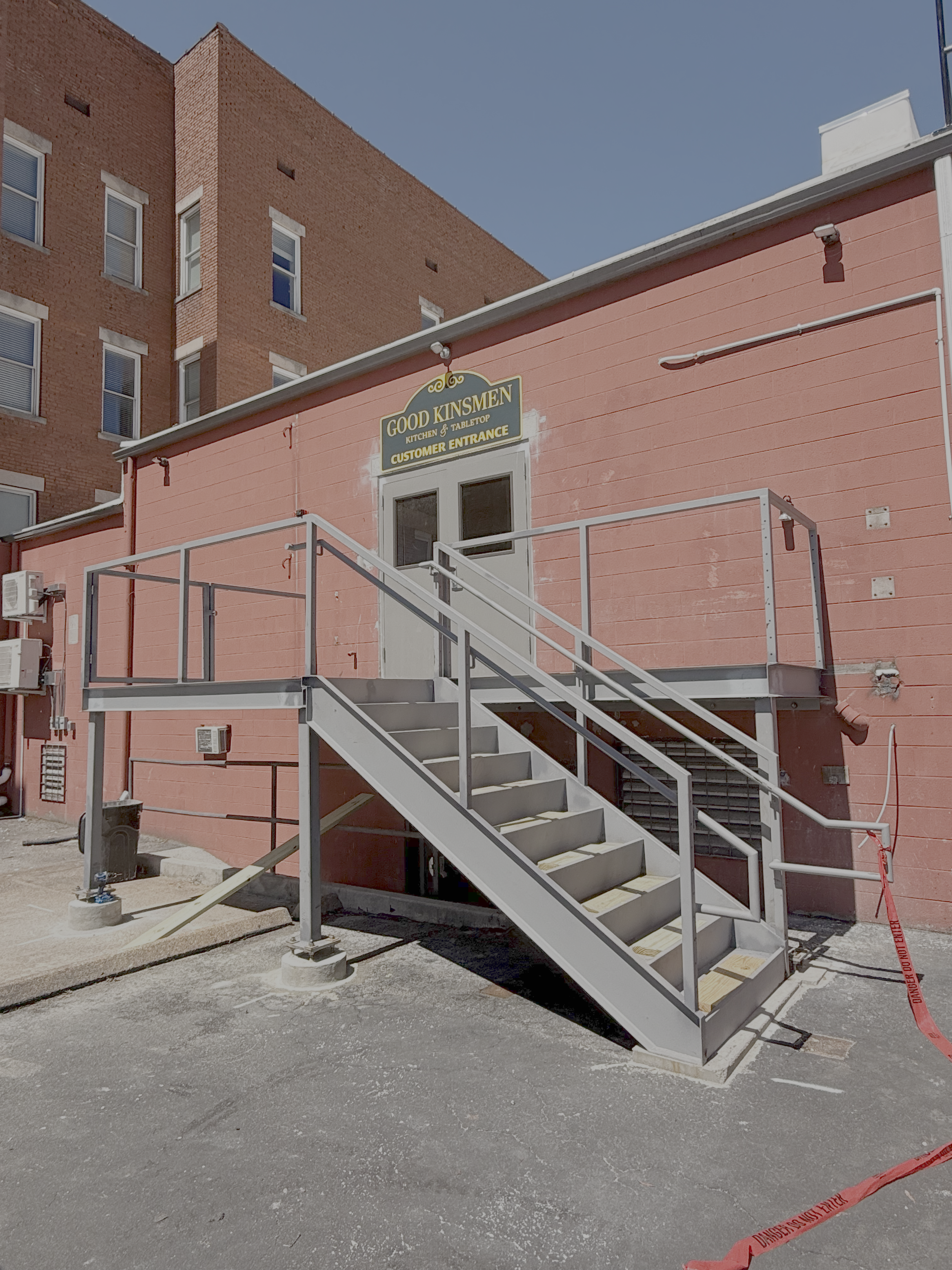 Metal outdoor staircase leading to a door on a pink building, with a green sign that reads 'Good Kinsmen Kitchen & Tabletop' and 'Customer Entrance'. The area is fenced off with a red caution tape on the ground.