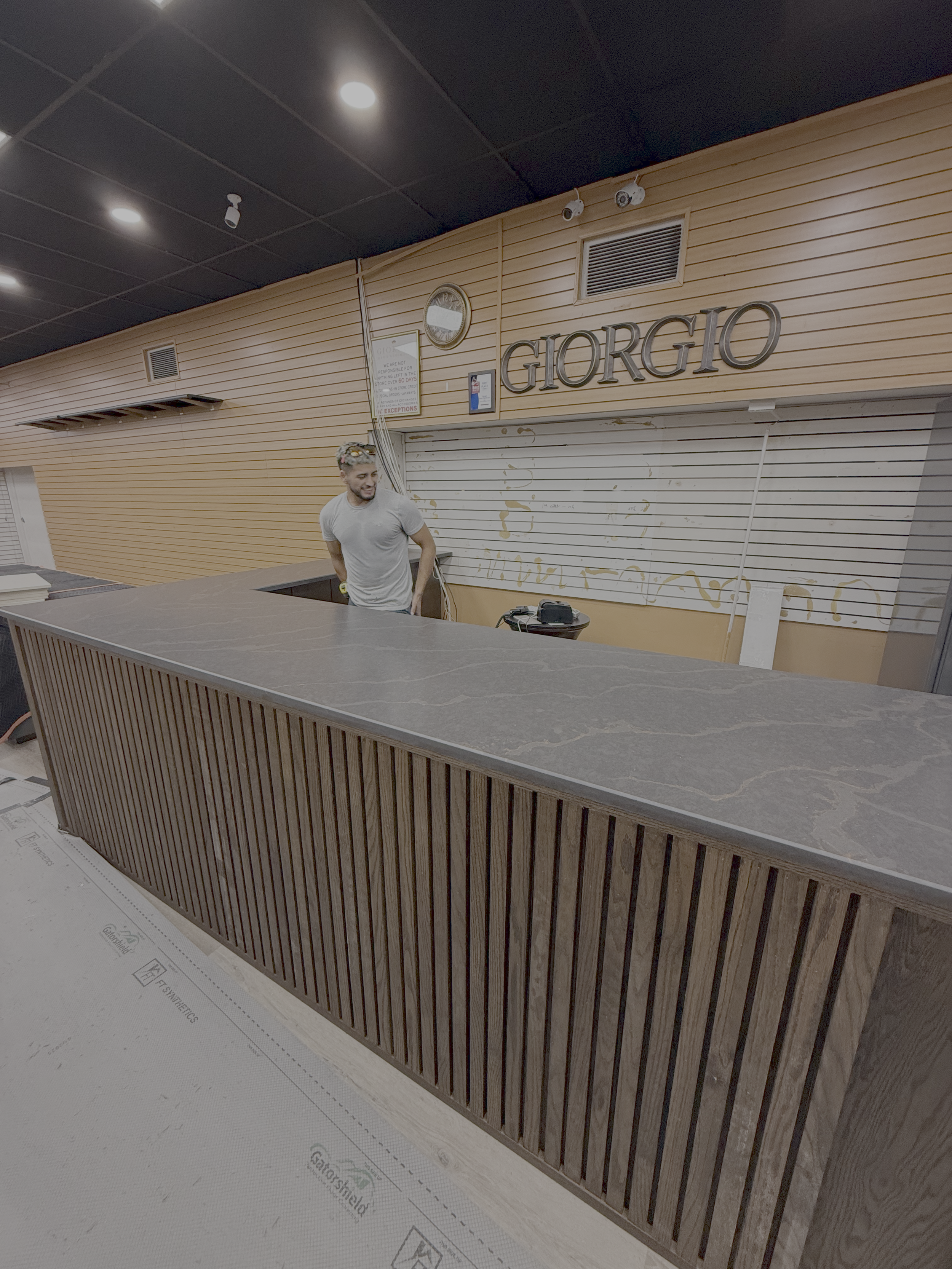 Interior of a store or office with a wooden slat front reception desk. A man stands behind the desk smiling. The background has wooden paneling and a sign that reads 'Giorgio'. There is a clock and framed items on the wall, and the ceiling has black 