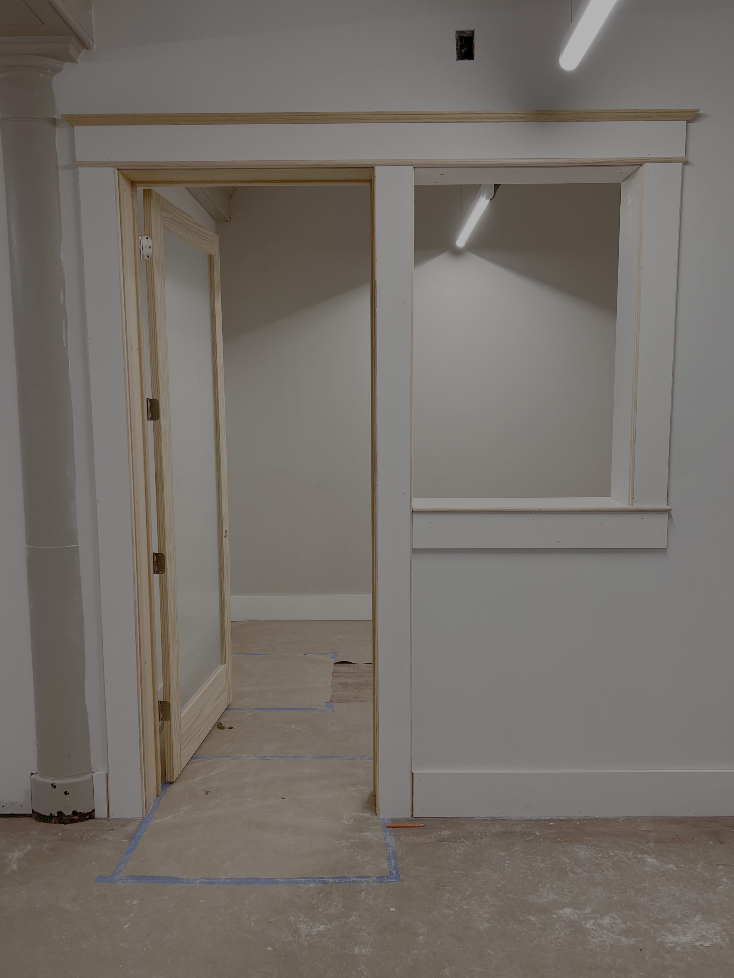 A partially constructed room with drywall framing, a doorway with a wooden frame, and a window frame on the right side. The floor is covered with protective paper and blue painter's tape, and the ceiling has exposed fixtures and a fluorescent light.