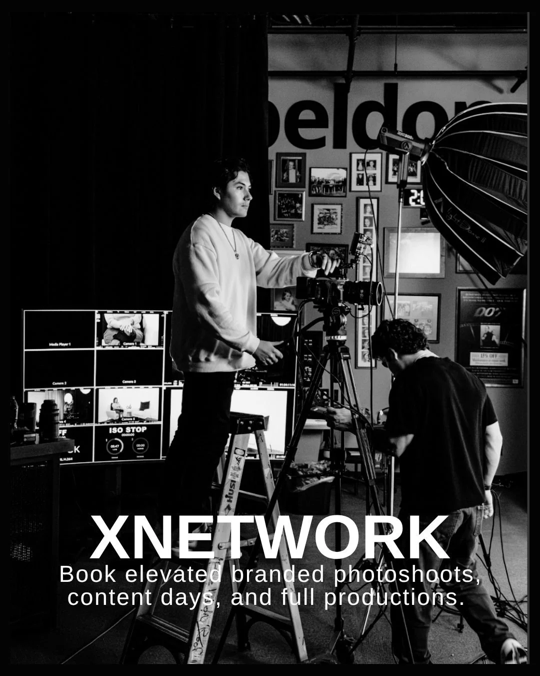 XNetwork Black and white photo of a woman standing on a ladder operating camera equipment during a photoshoot, with a man adjusting equipment nearby in a studio with framed photos and a partial word "Welcome" in the background.