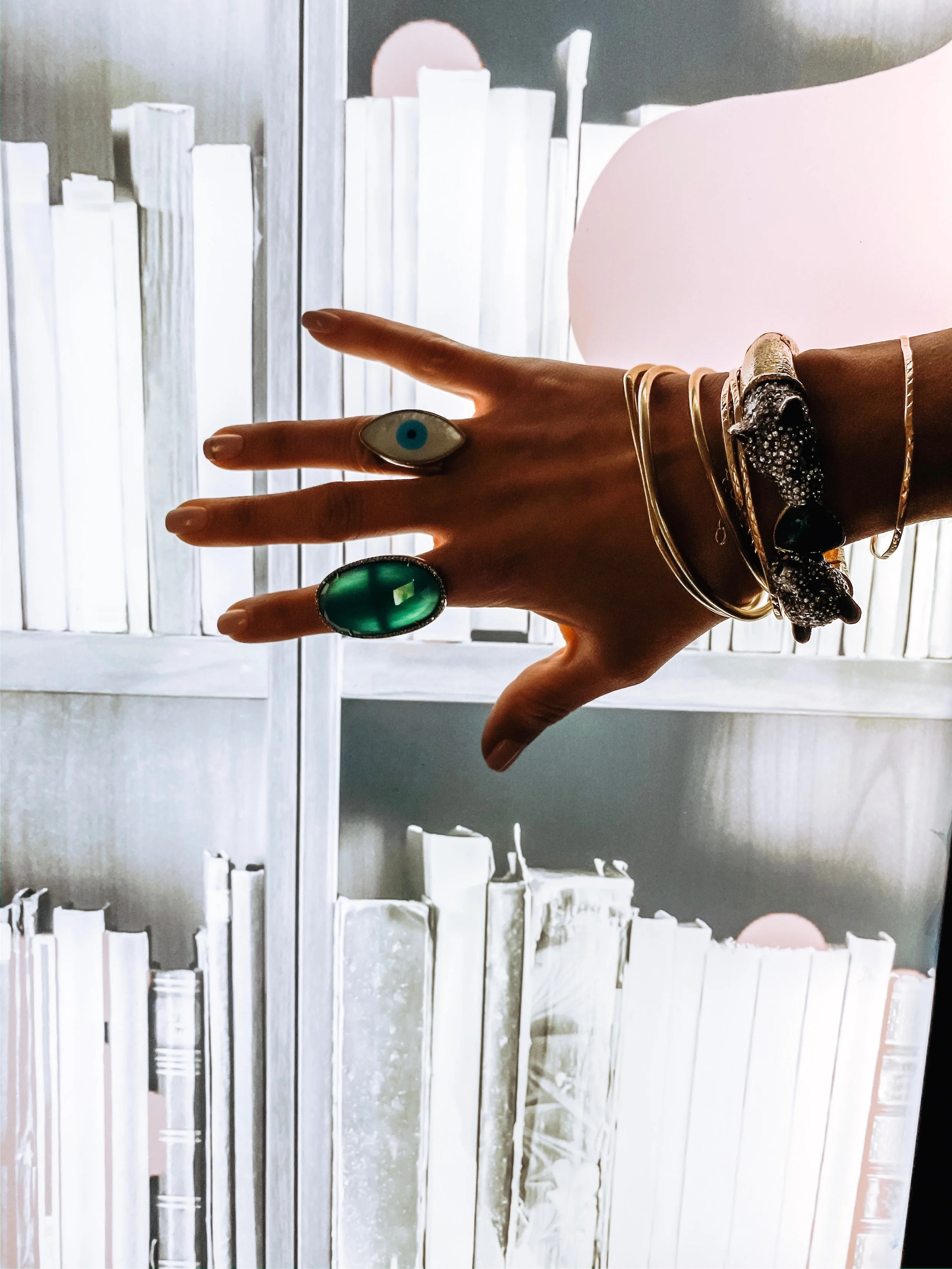 Xitlalt's hand with multiple rings and bracelets, including a ring with an eye design, extended in front of a window with white blinds, with the person's face not visible.