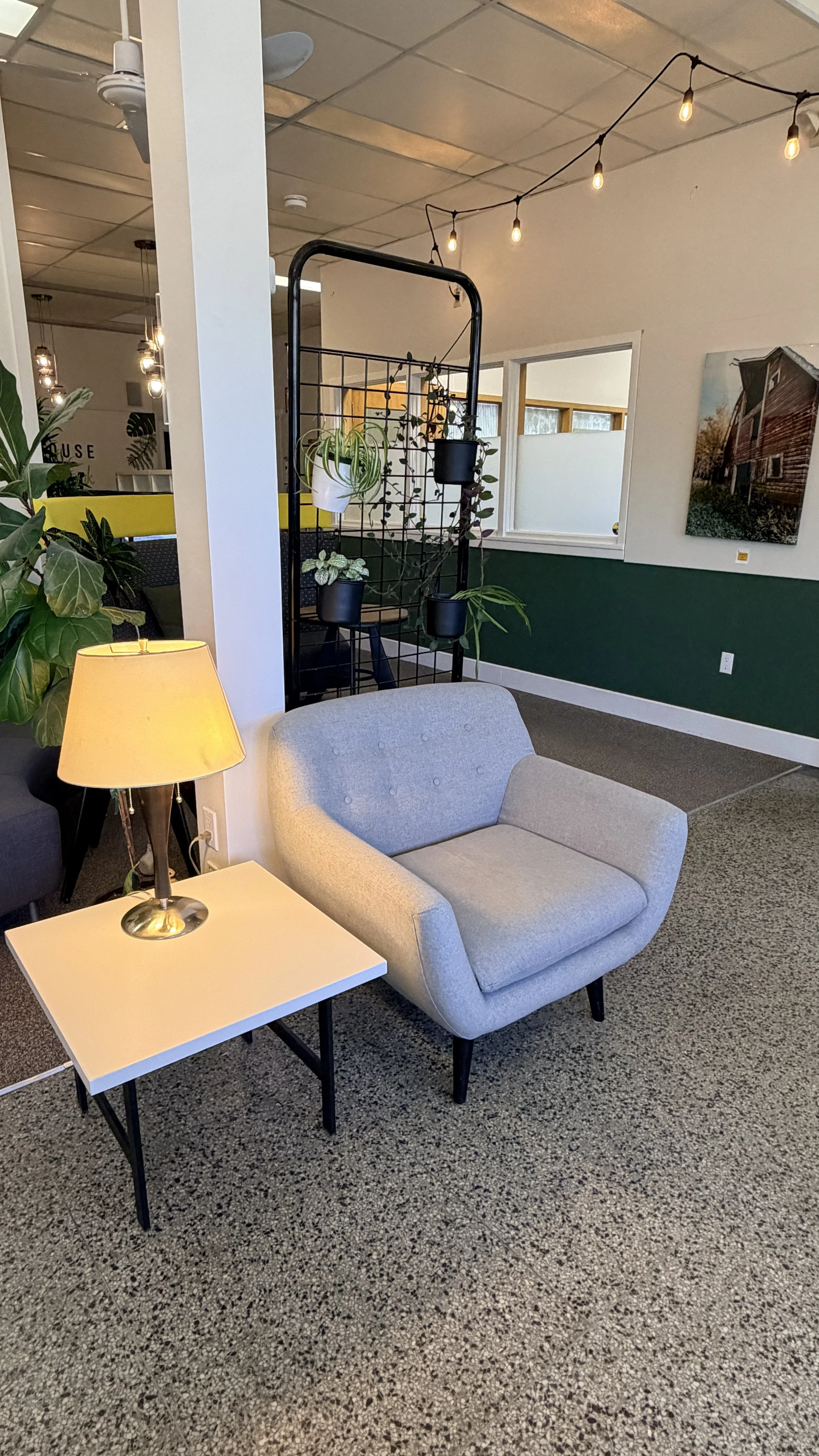 A cozy indoor space with a gray armchair beside a white side table with a lamp, and a collection of hanging potted plants on a black metal stand, with string lights overhead and artwork on the walls.