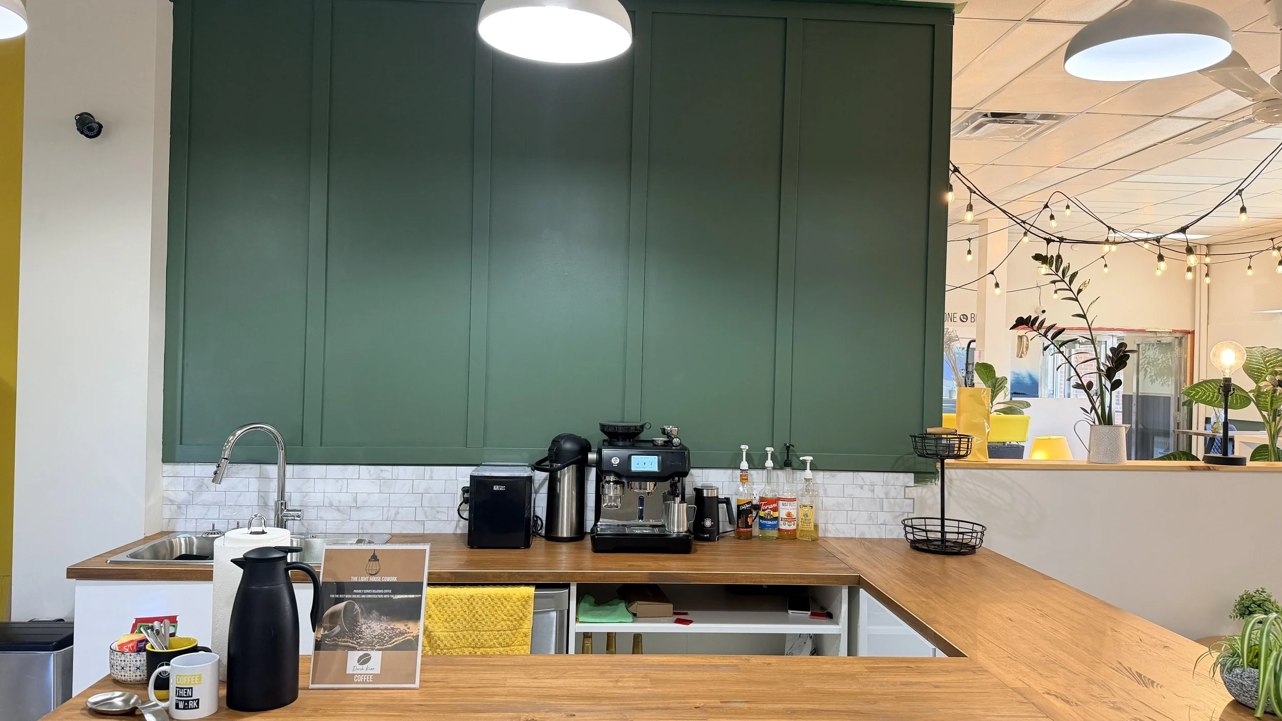 Coffee station in a café with coffee machines, brewing supplies, and decorative plants, with hanging string lights and a green wall.