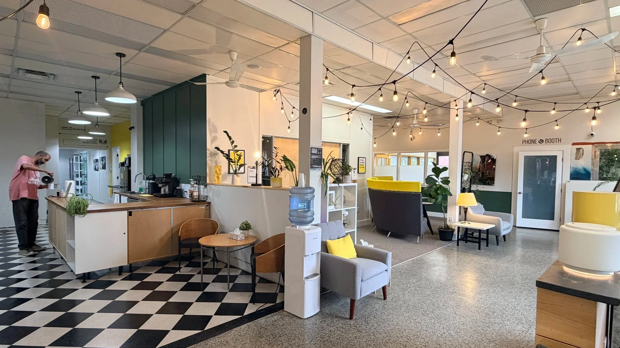 Indoor cafe or lounge with modern decor, hanging string lights, potted plants, seating area with armchairs and couches, and a person pouring a drink behind the counter.