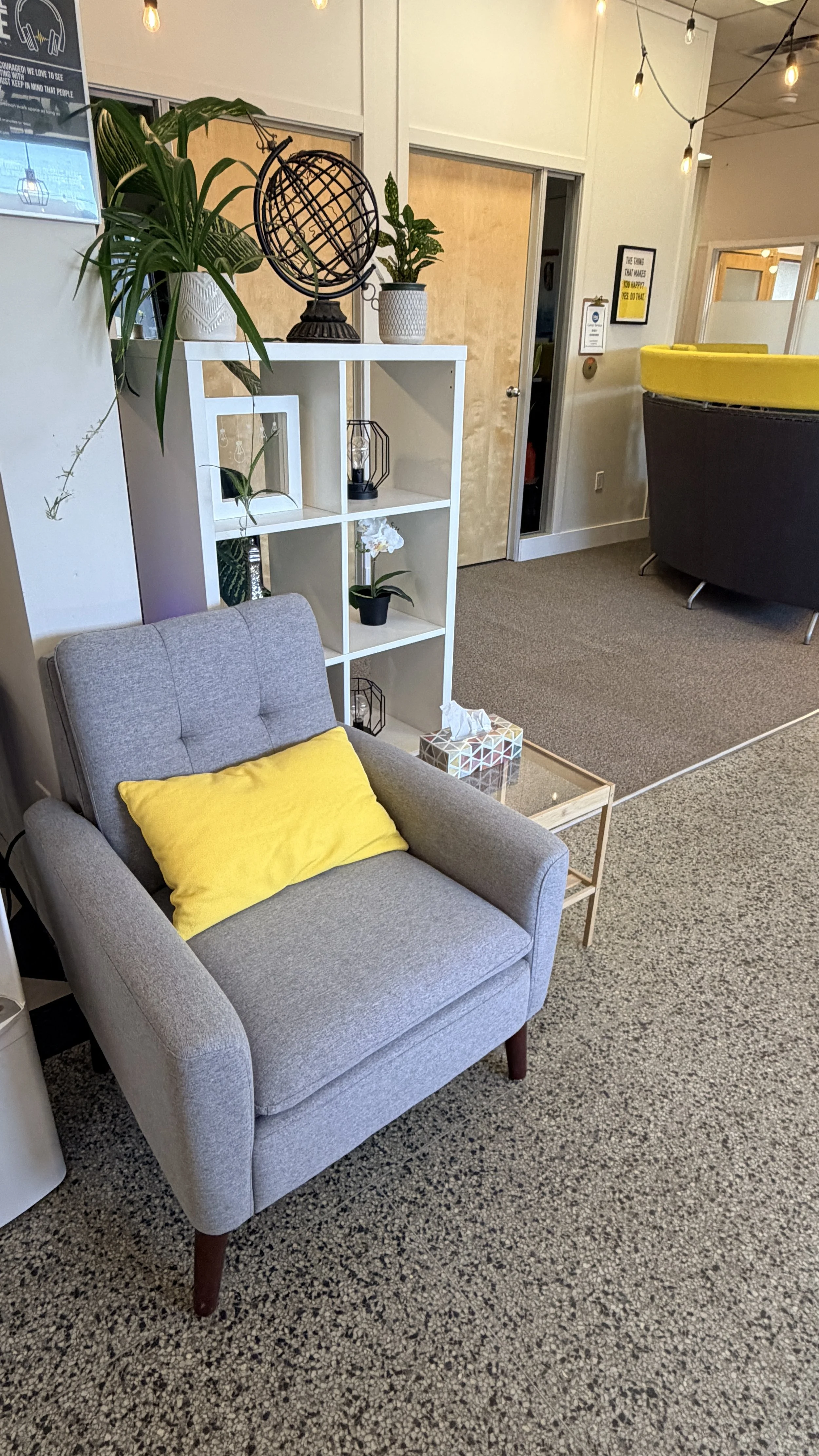 A cozy waiting area with a light gray armchair that has a yellow pillow on it, a small glass-top side table with a tissue box, a white shelving unit with plants and decorative items, and a carpeted floor. There are string lights hanging from the ceil