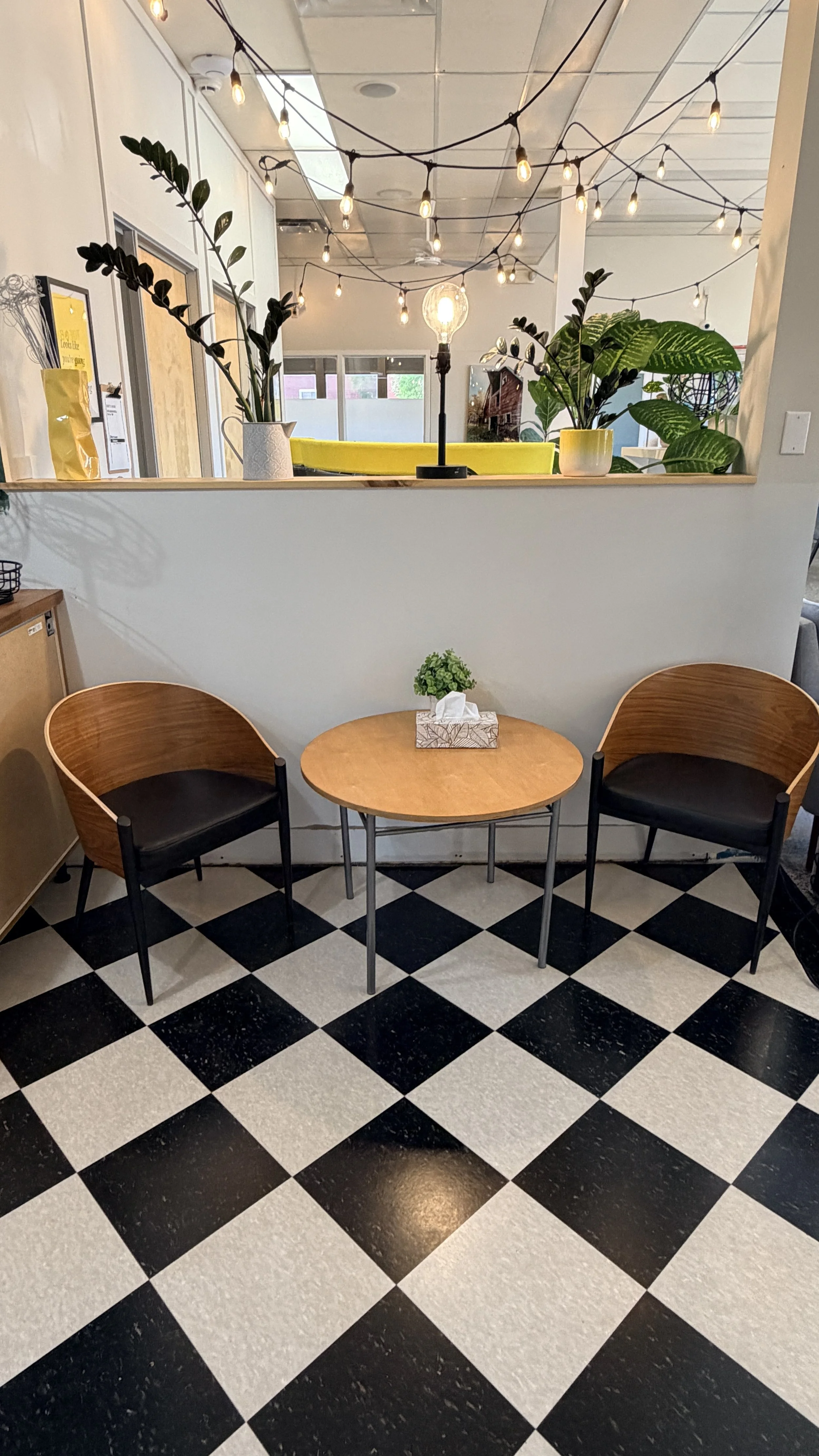 A cozy indoor seating area with two wooden chairs with black cushions, a small round wooden table with a plant and tissue box, a black and white checkered floor, decorative string lights, and plants on a ledge overlooking an open space with additiona