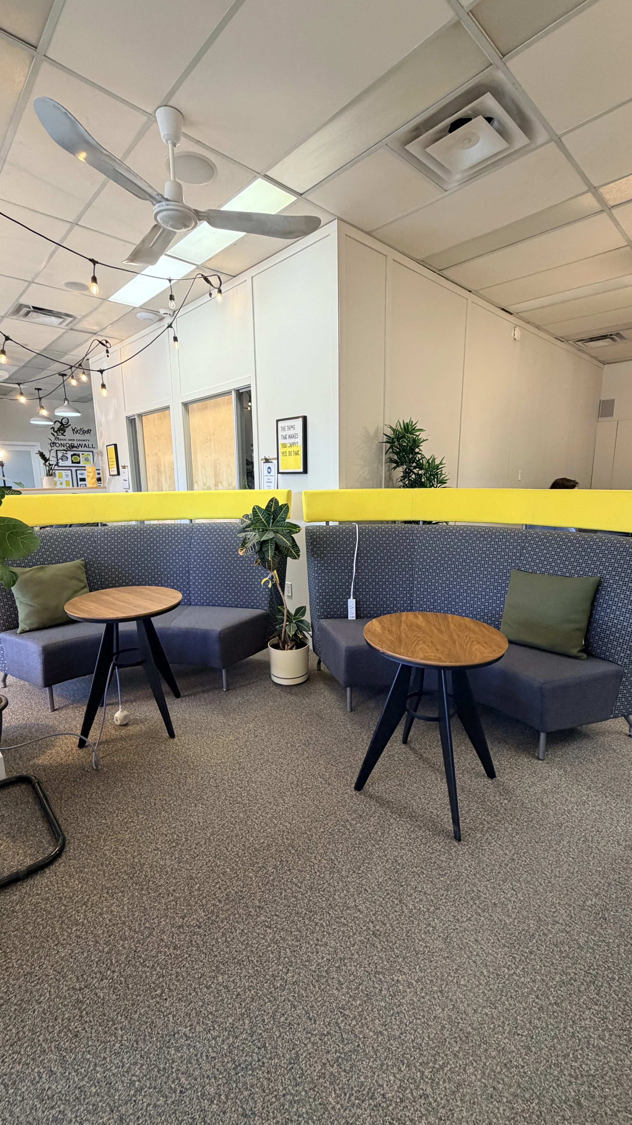 Indoor lounge area with two gray sofas, each with a green pillow, two small round wooden tables, potted plants, string lights, ceiling fan, and ceiling vents. In the background, there are windows and a door with signs on the walls.