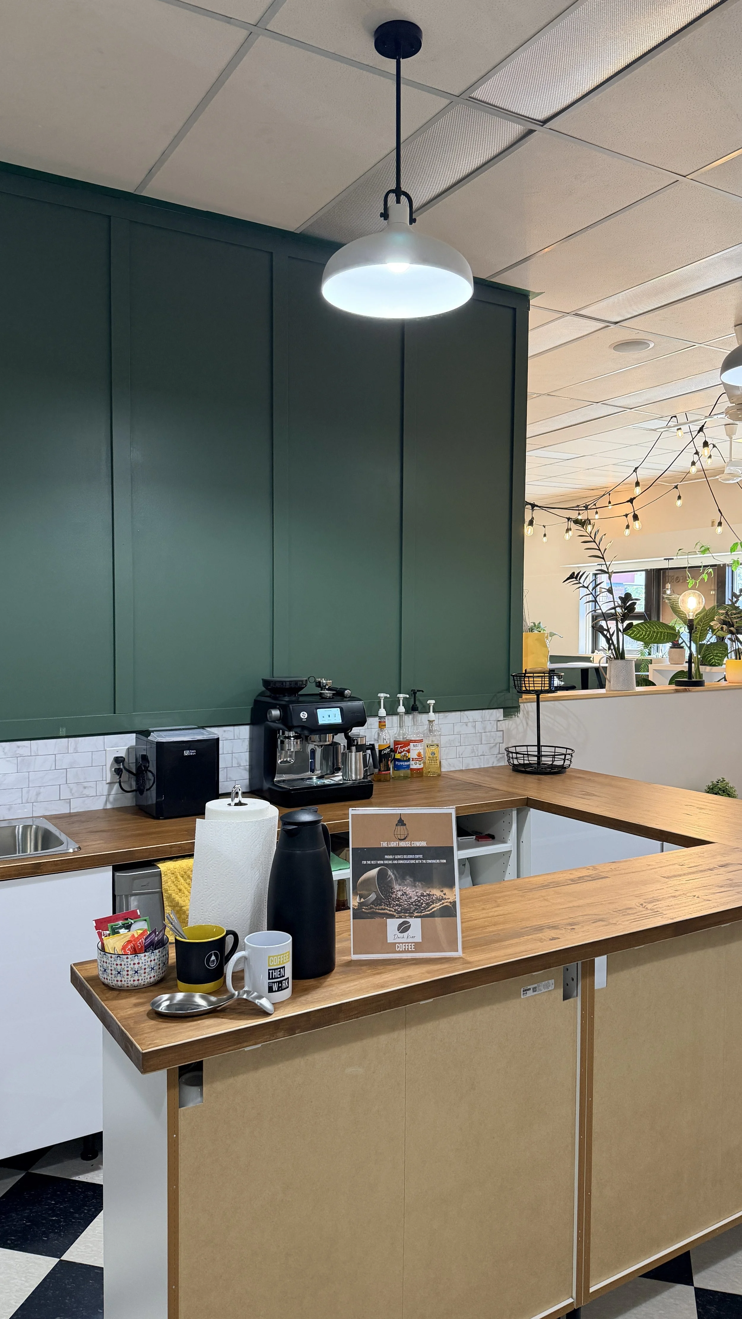 A coffee bar with a wooden countertop, coffee-making equipment, and cups, set against a dark green wall with plants and light strings in the background.
