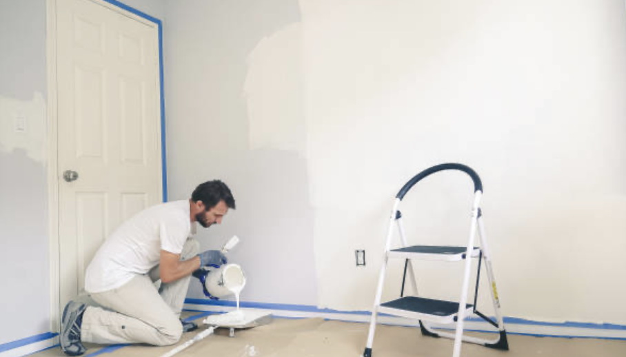 A man painting a wall in a room with paint roller, ladder nearby, door in background.
