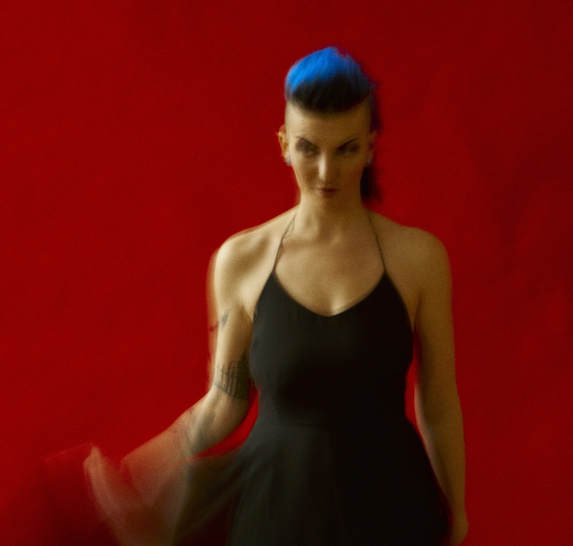 A person with blue hair wearing a black halter dress stands against a red background. The image is slightly blurred.