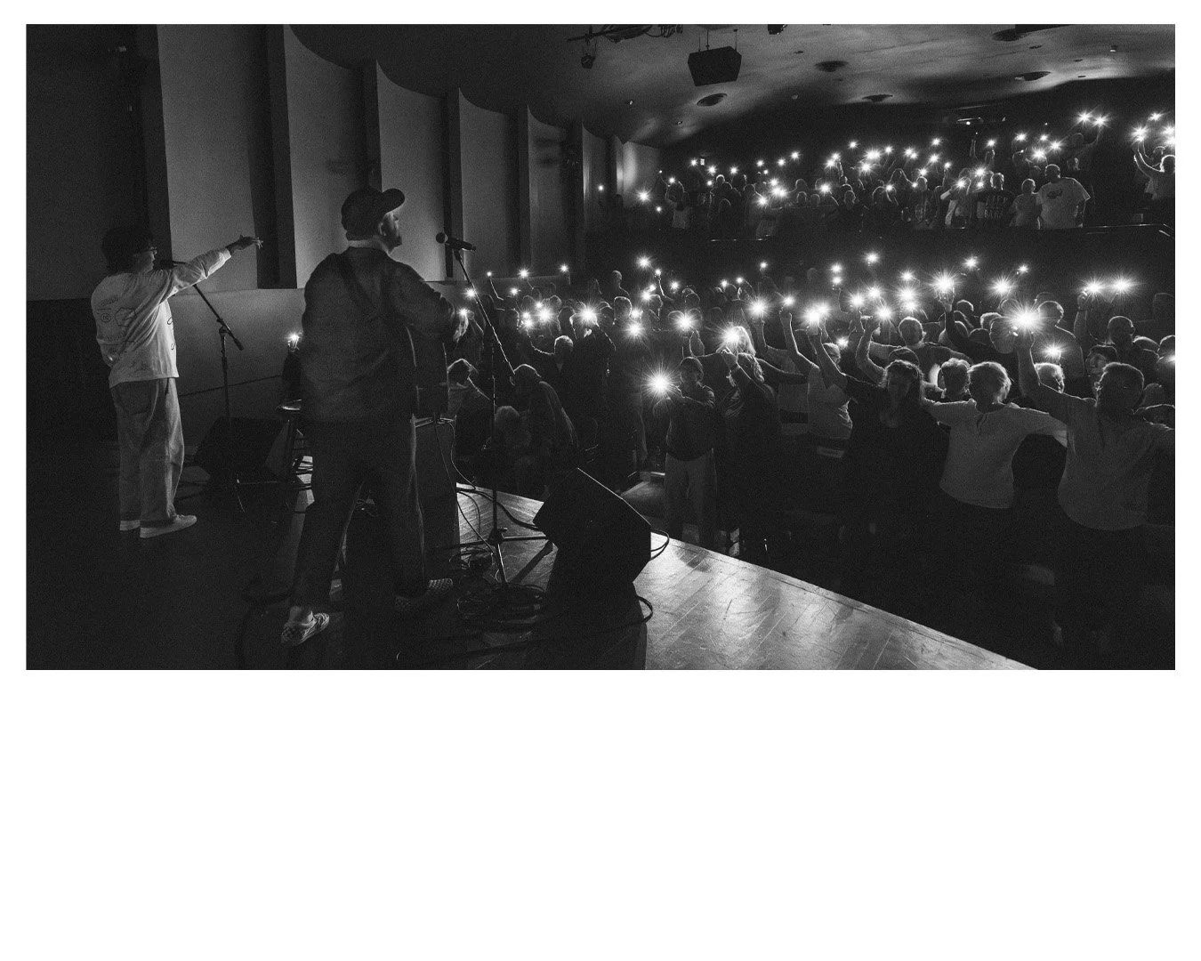Black and white photo of a live performance with two musicians on stage and an audience holding up lit smartphones.