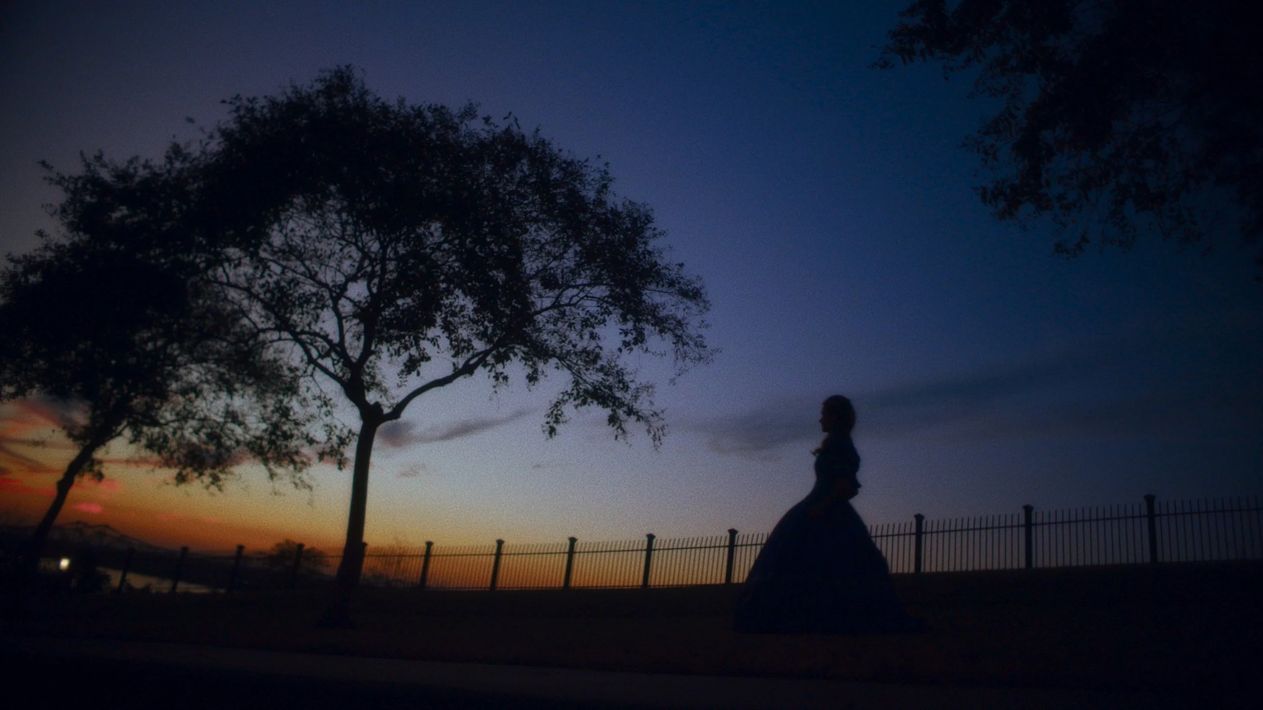 A silhouetted person in a hoop skirt walks along a fence