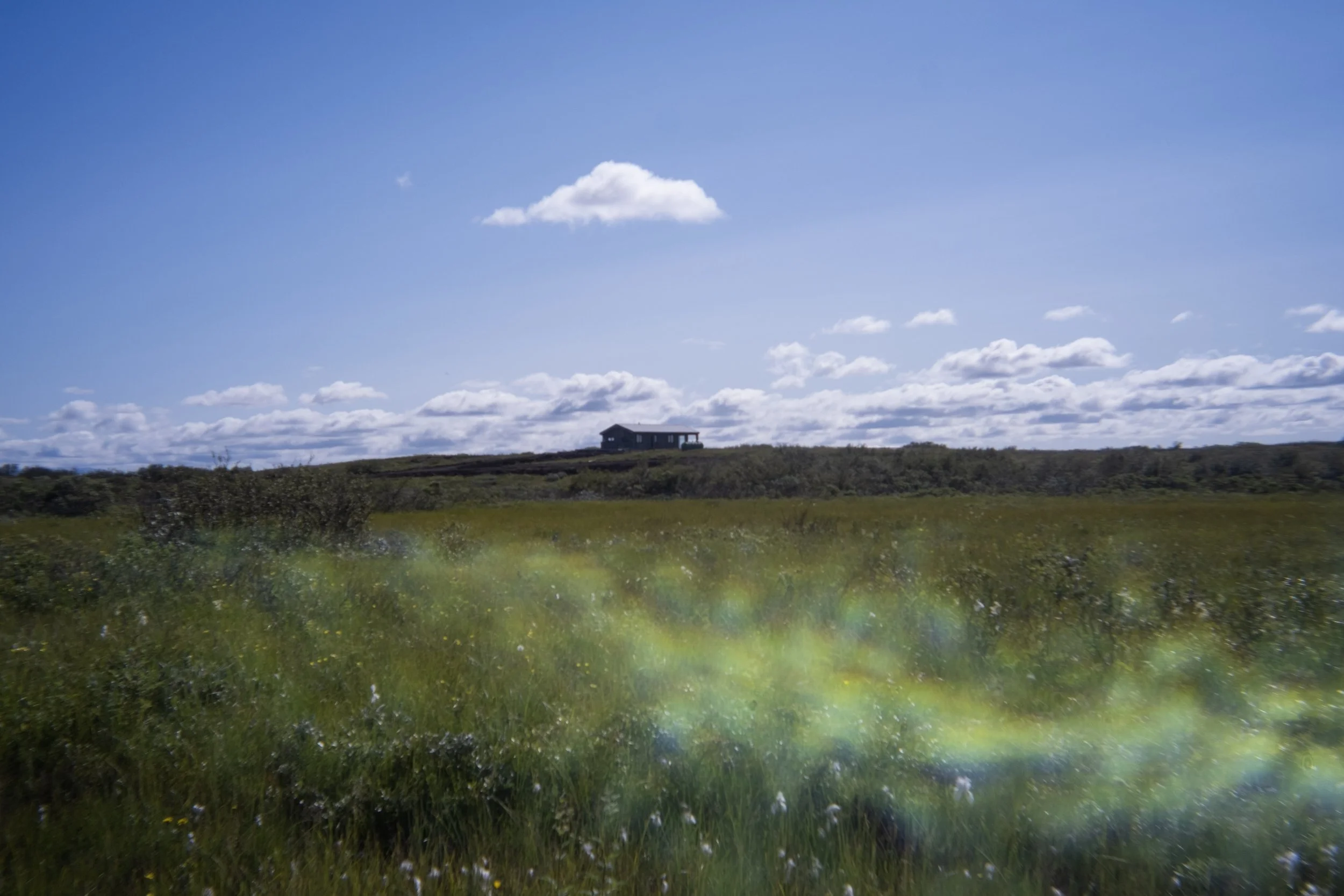 Cabin sit in the distance amongst clouds and above the grass with a rainbow sun glare in Iceland