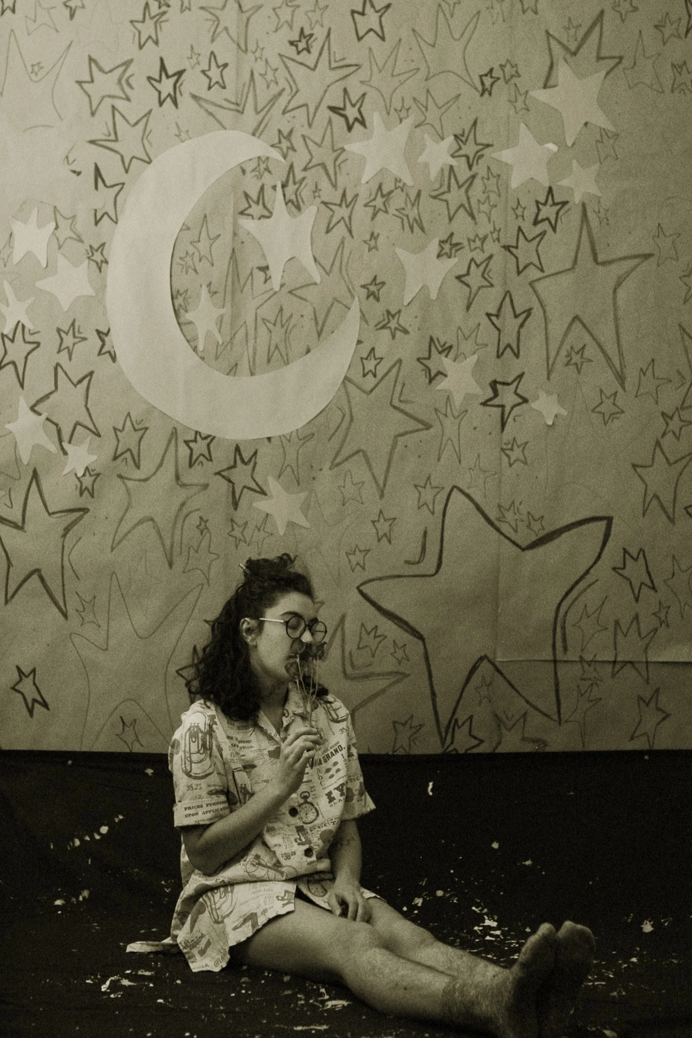 a start covered brown paper backdrop with a paper moon and stars cuts off at the bottom into a splattered cloth. margo santiago sits legs outstretched in a short newspaper dress holding flowers up to her noses in deep inhale. she wears glasses.