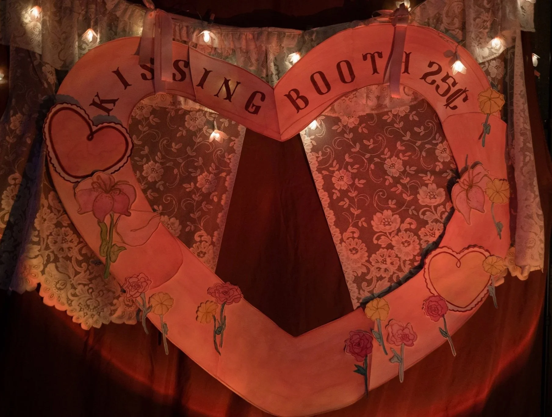 a red backdrop with a doiley half curtain sit behind a heart shaped water color backdrop reading 'KISSING BOOTH' there are water color hearts plus pink and yellow flowers with stems.