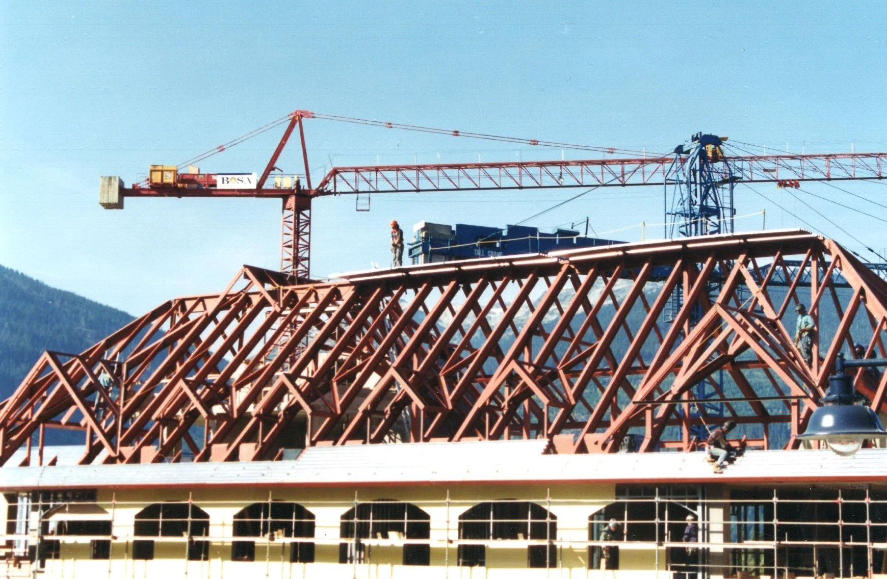 Construction workers assembling a steel framework on top of a building, with a large crane in the background against a mountainous landscape.