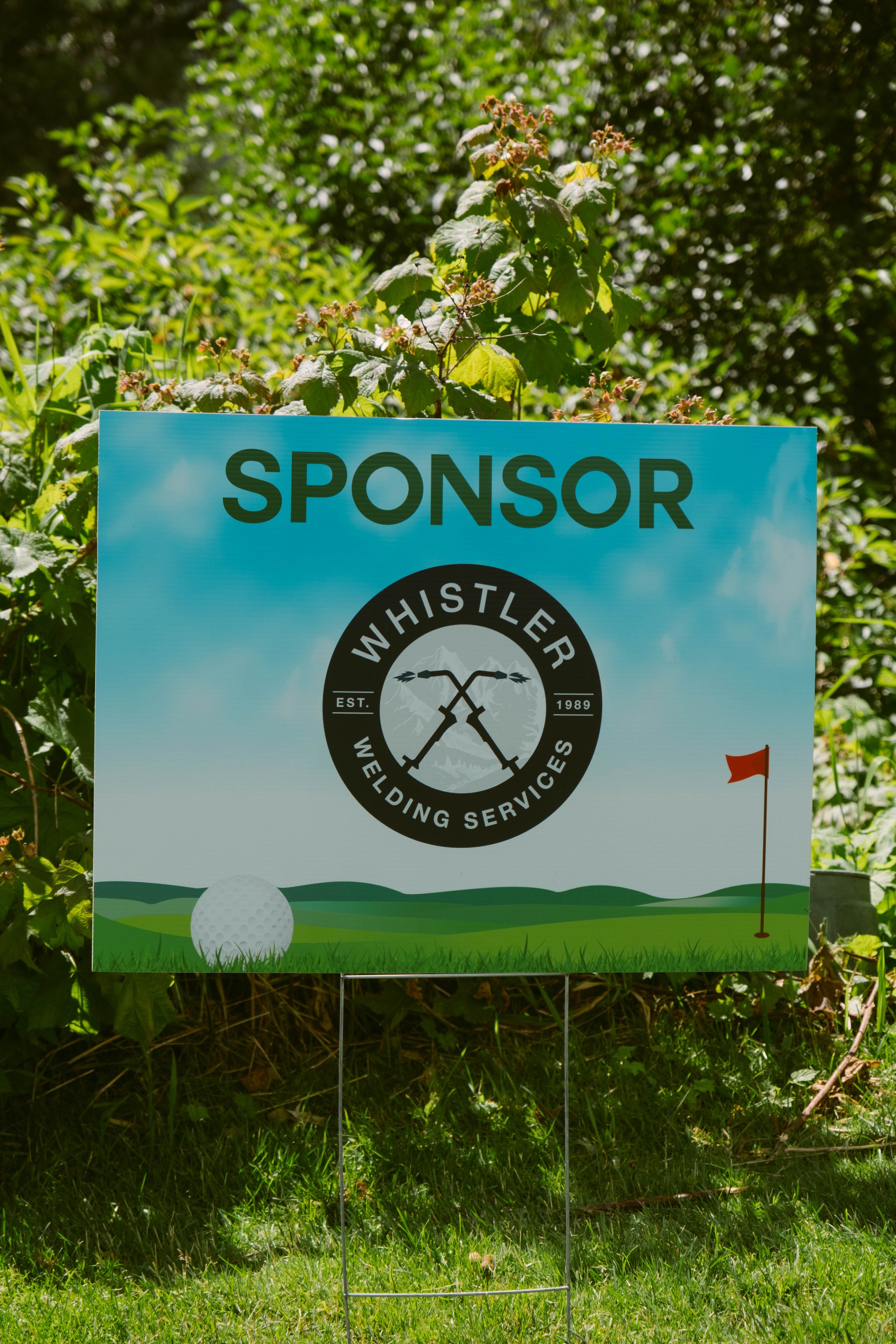 A sign for Whistler Wedding Services with a golf course illustration, a golf ball, and a flag on the green, set outdoors among green plants and bushes.