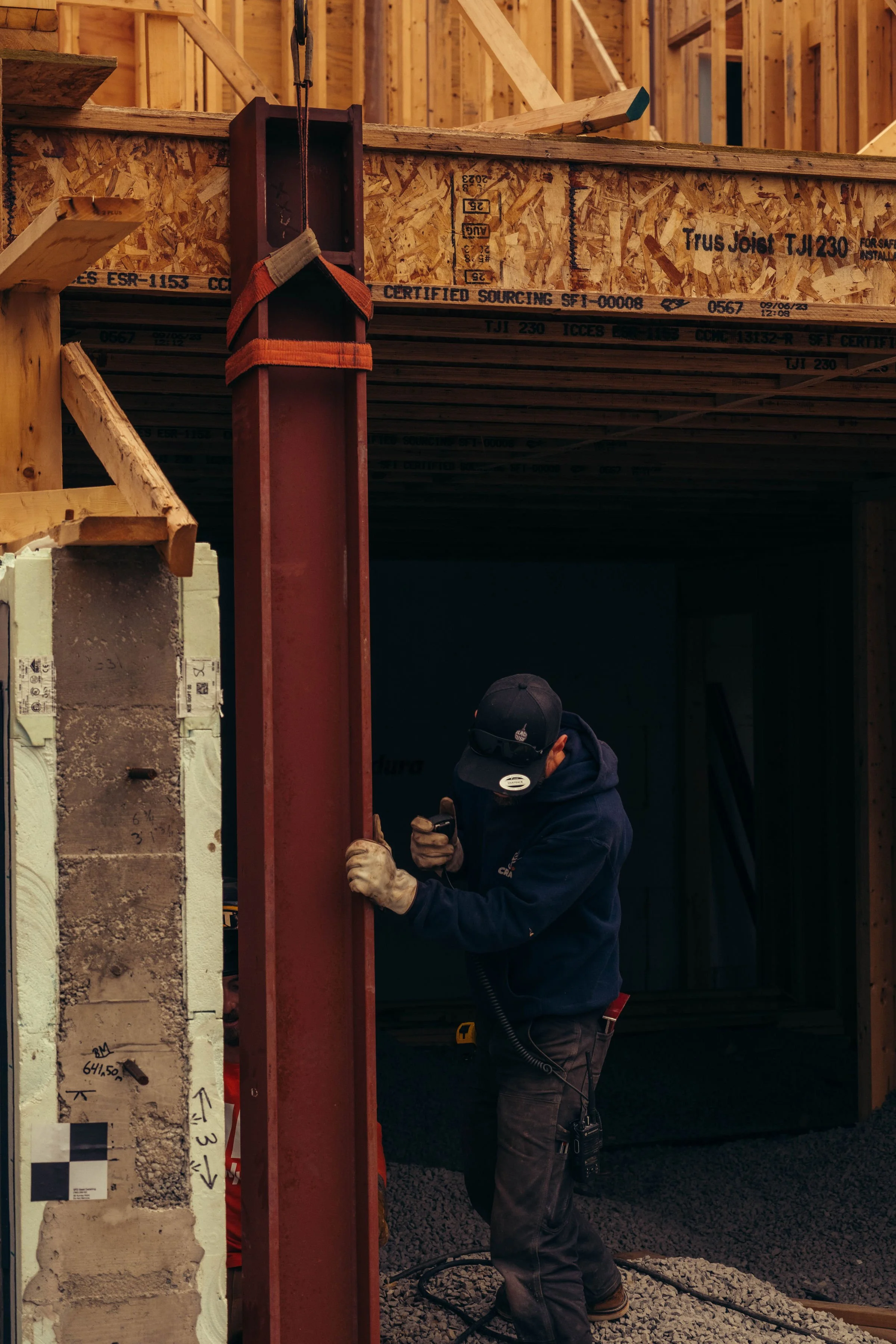 A construction worker wearing a black cap, safety gloves, and a navy hoodie is working on a construction site. He is securing a large red steel beam to a vertical support. The background shows an unfinished wooden frame of a building under construction with concrete blocks and wood studs visible.