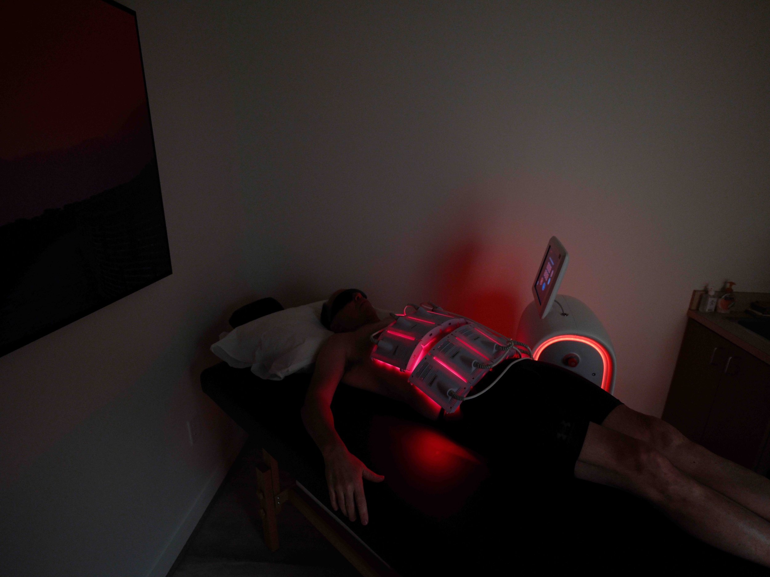 A person lying on a treatment table undergoing a red light therapy session. The individual is wearing protective goggles and has a device emitting red light placed on their torso. The room is dimly lit with a display screen visible in the background.