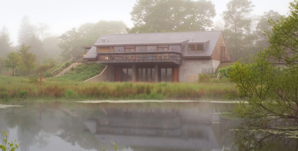 mist-shrouded, modern wood and concrete building nestled into a grassy hillside. Its reflection and the surrounding fog-covered trees are captured in the still water of a pond in the foreground.