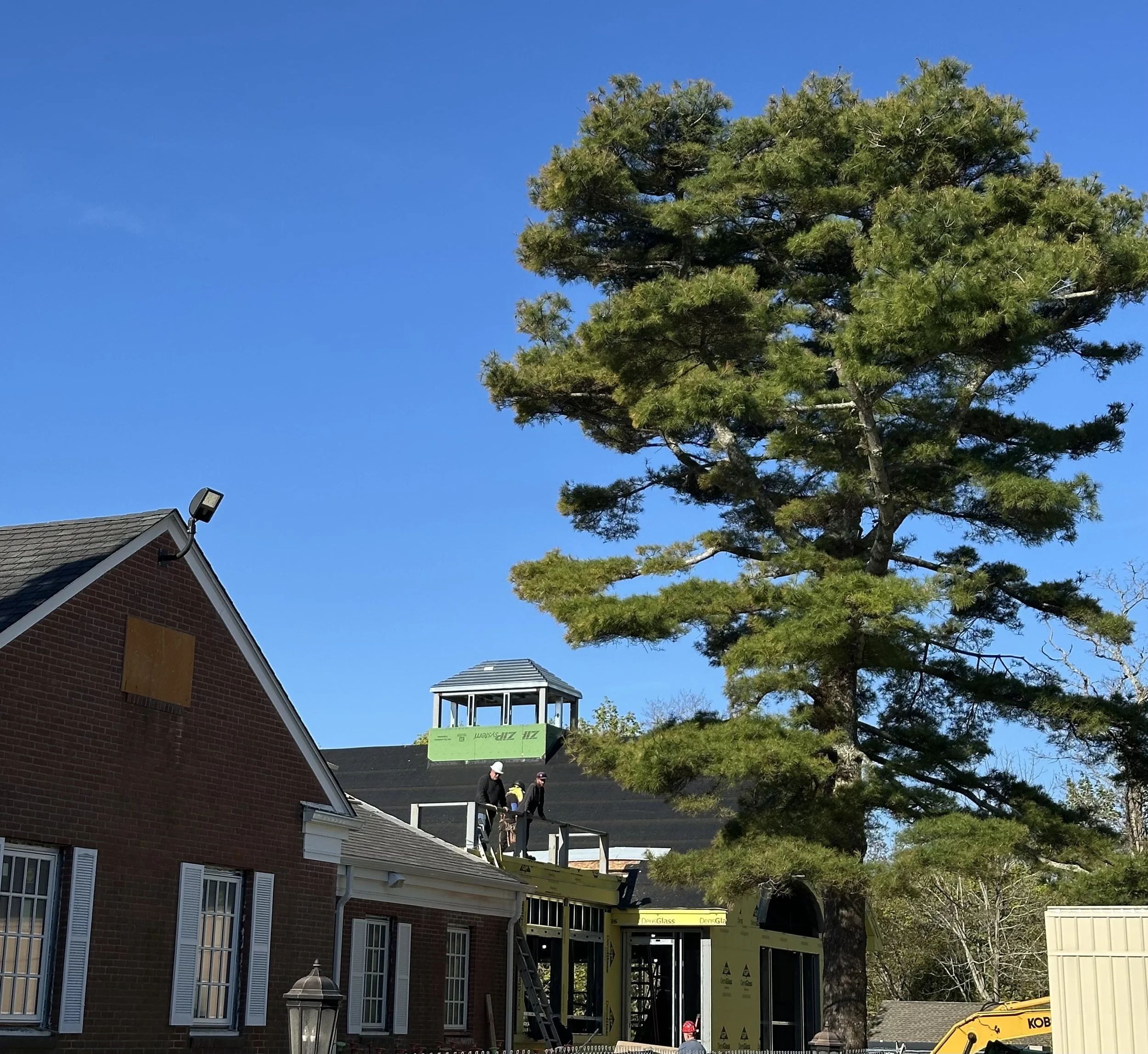 In this updated view of the Shelter Island Library project, construction has progressed significantly, showing the new addition being integrated with the existing brick building. Two workers in hard hats are visible on a high platform of the addition