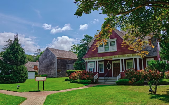 bright, sunny daytime photograph of a historic museum complex featuring a two-story red building with a white porch and shingled upper roof, situated on a manicured green lawn alongside weathered grey shingle outbuildings.
