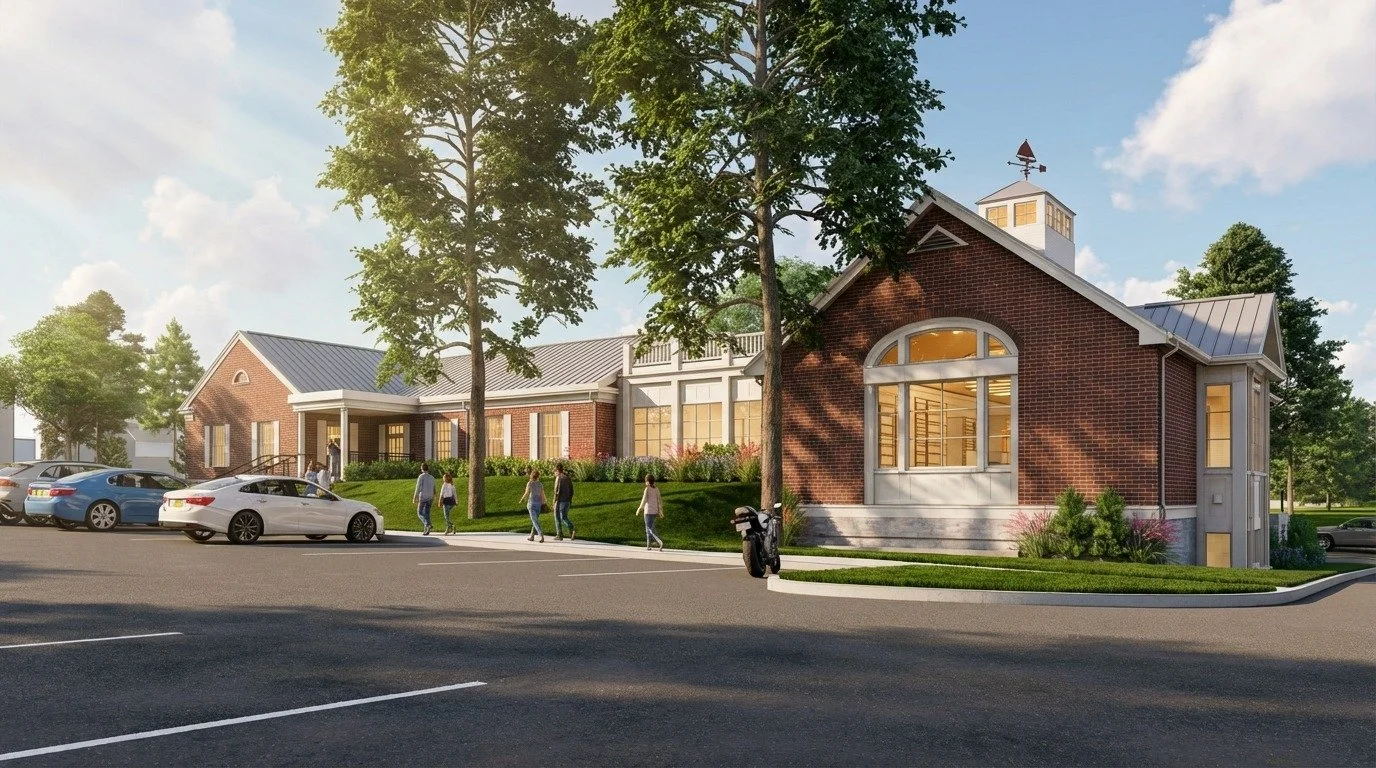 A red brick library exterior with a gray metal roof and a small white cupola on top. It features large arched windows, a green lawn with tall trees, and a parking lot in the foreground with a few cars and people walking toward the entrance.