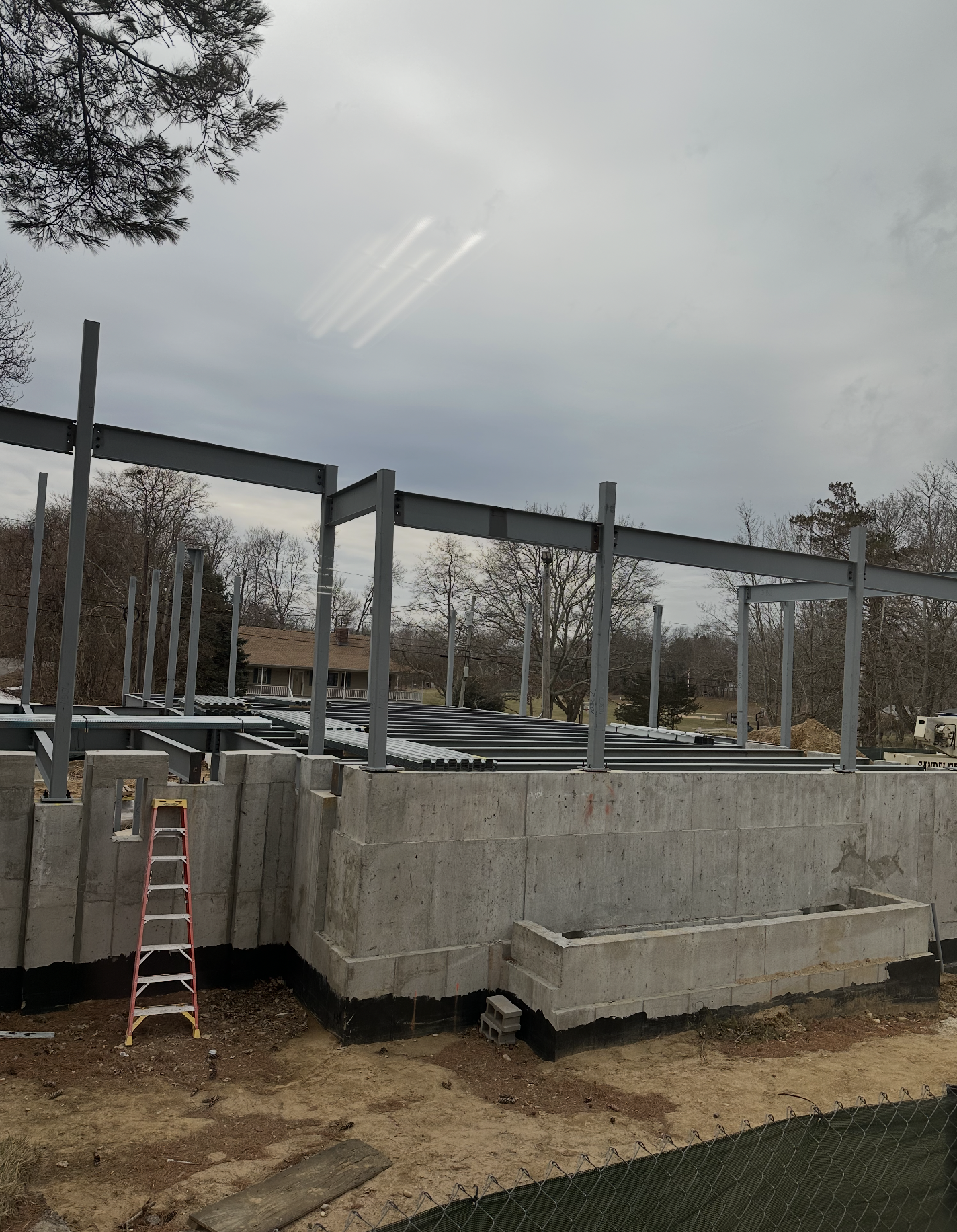 A wide-angle, eye-level photograph shows the structural steel framework and concrete foundation of a building under construction. Several vertical gray steel columns support horizontal beams, forming the skeleton of the new addition against a cloudy 