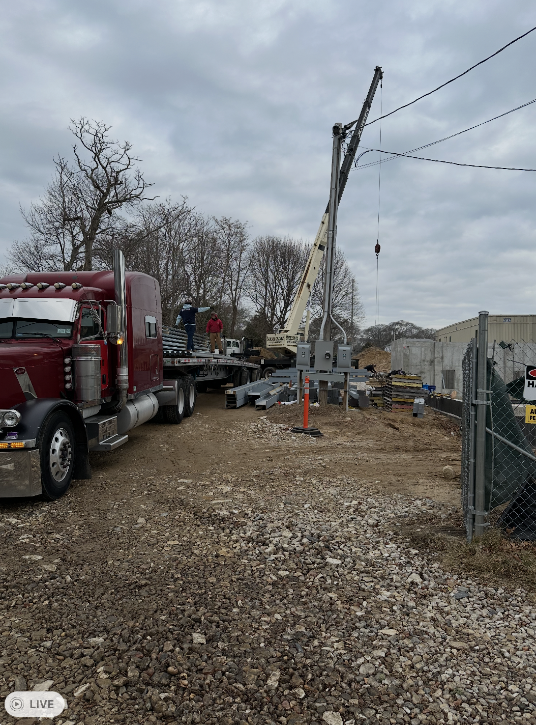 A maroon semi-truck with a flatbed trailer sits on a gravel and dirt construction site, loaded with gray steel beams. In the background, a tall crane stands near a concrete foundation, while two workers are visible on the trailer managing the materia