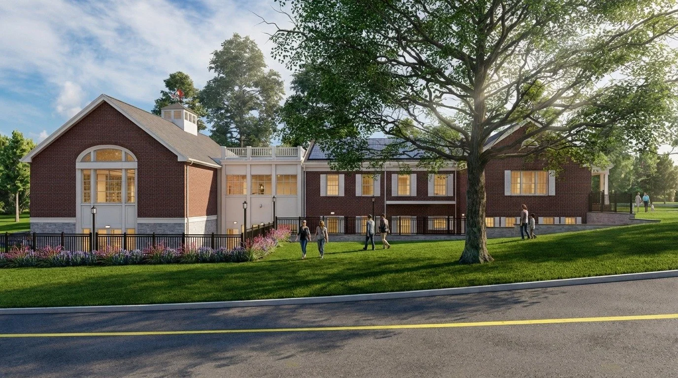 A long, red brick building with white trim and a gabled roof. People walk on a grassy slope in the foreground, bordered by a black metal fence and a paved road. A large, leafy tree stands to the right, and solar panels are visible on the roof.