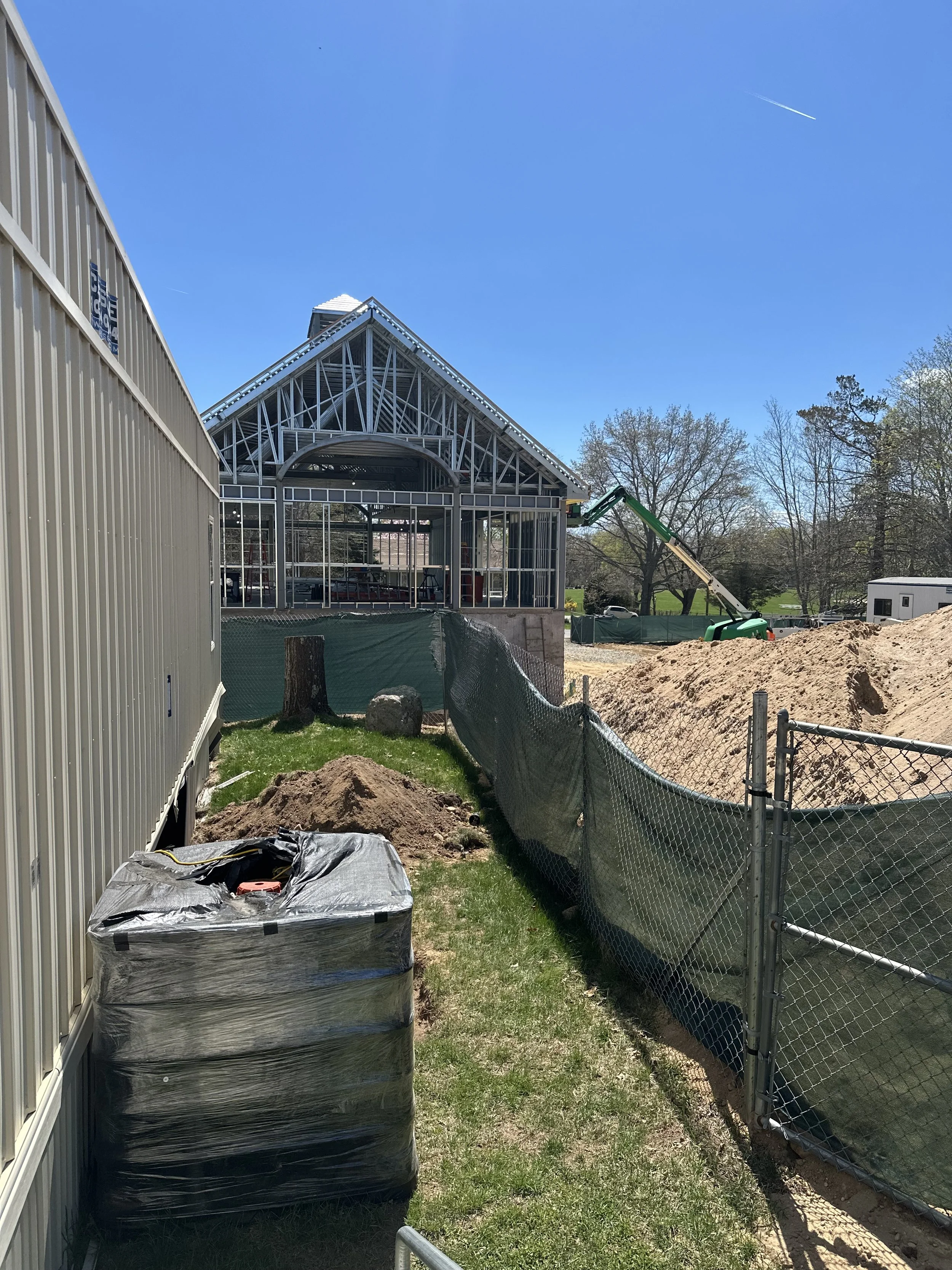 The image shows the steel-framed skeleton of the new Shelter Island Library addition rising under a clear blue sky. The structure features a high-peaked gabled roof and a distinct arched entrance, currently consisting only of silver metal studs witho